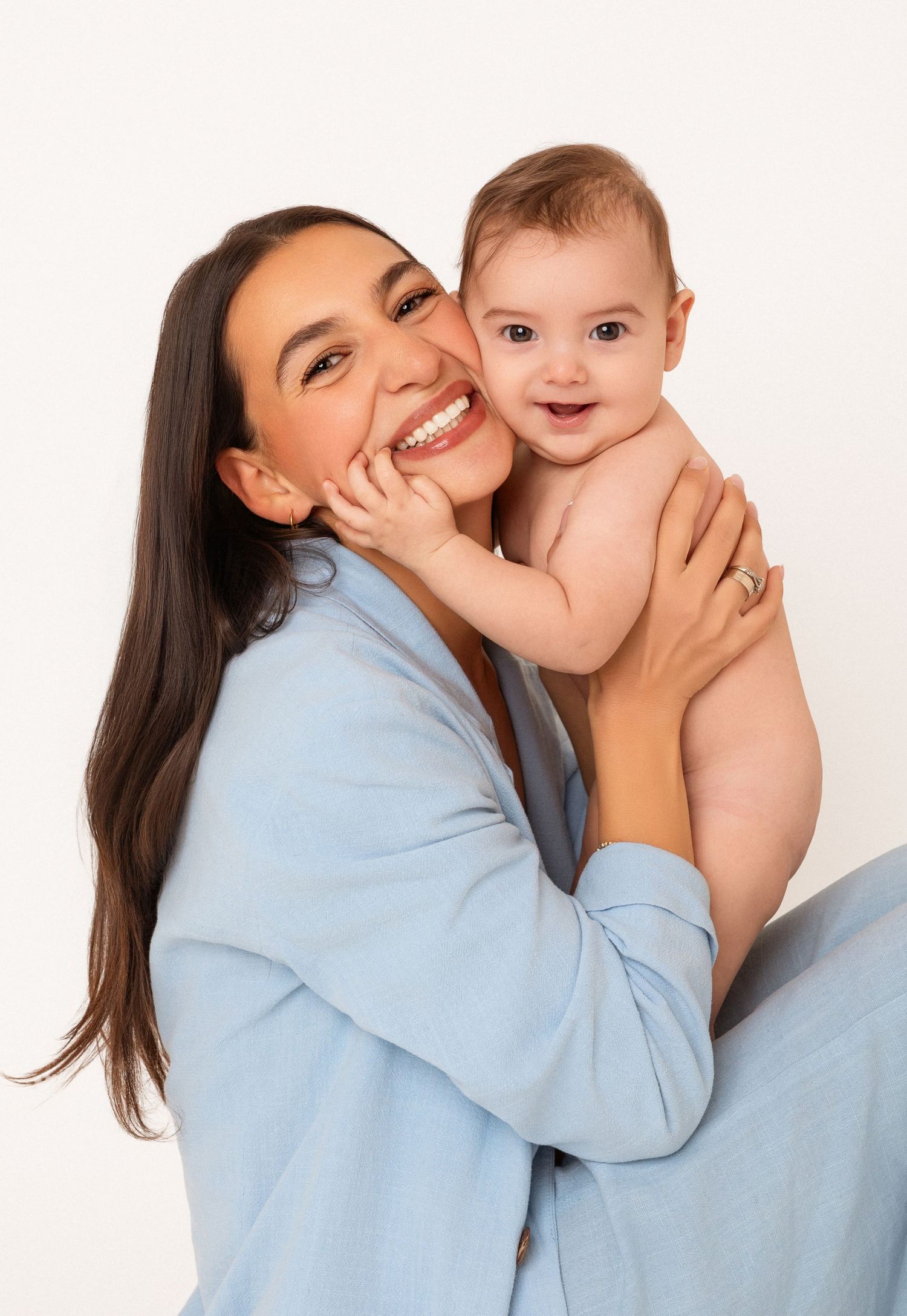 Smiling mother with long dark hair holding cheerful baby in blue cardigan.