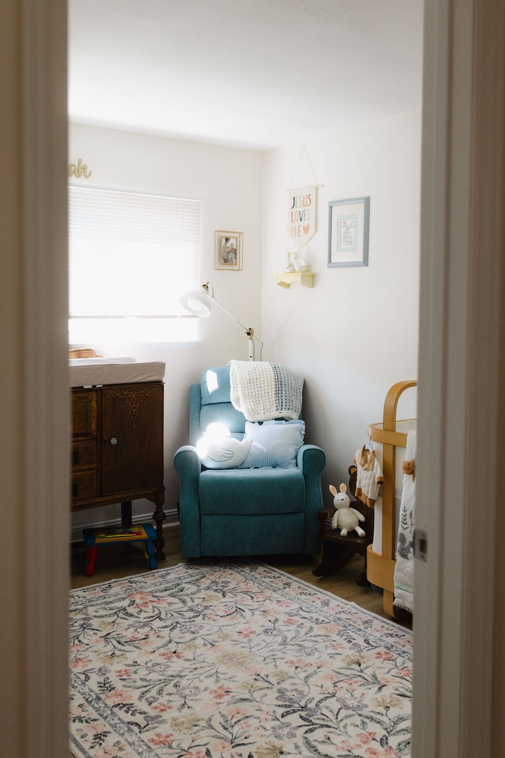 Bright nursery room with teal armchair, floral carpet, and white walls viewed through doorway.