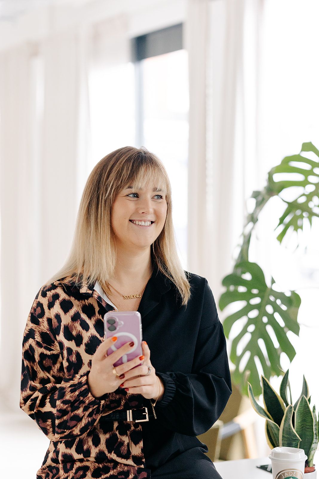 Two people in casual attire smile while holding phones and sitting in bright office space with houseplants.