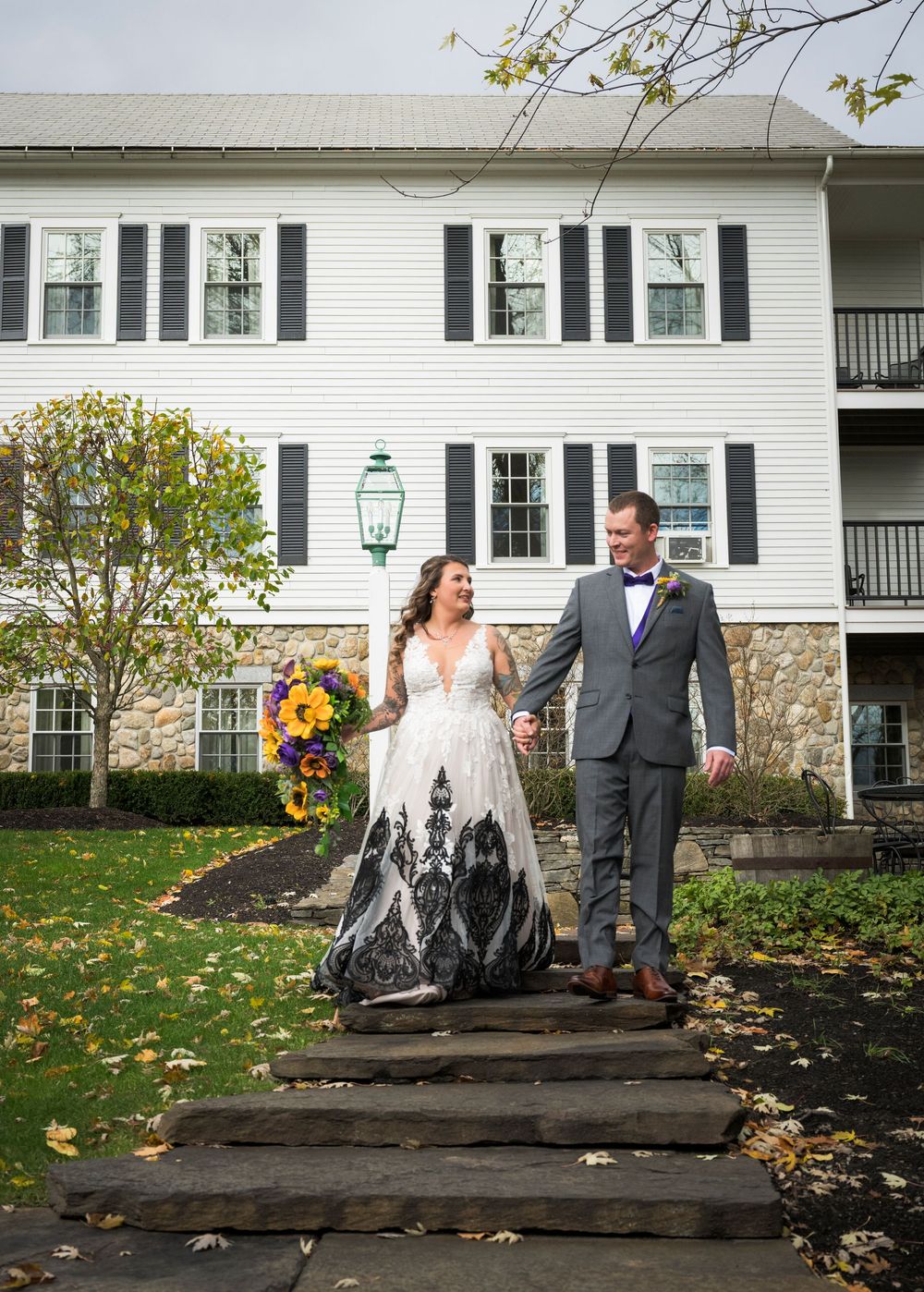A couple takes romantic photos in front of a white house with stone foundation on a fall day.