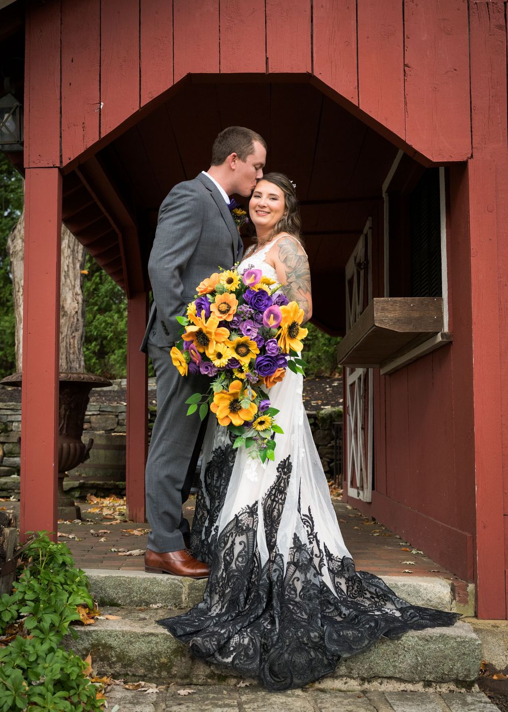 Newlywed couple embraces in front of red barn with vibrant sunflower bouquet and black and white wedding dress.