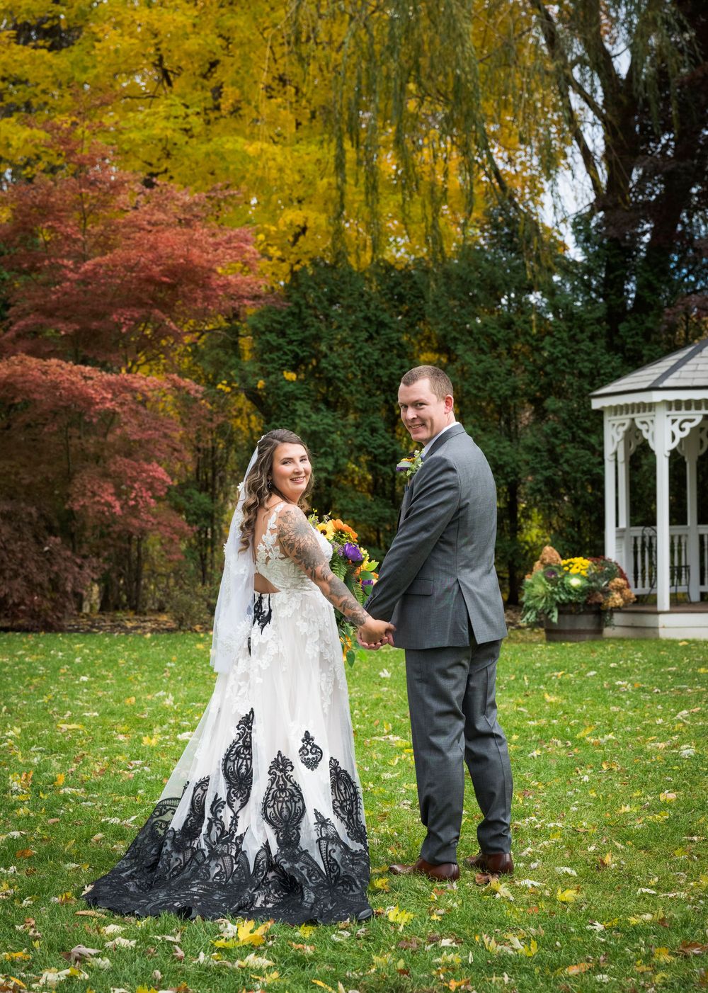 A wedding couple poses on a lawn with autumn foliage and a white gazebo in the background.