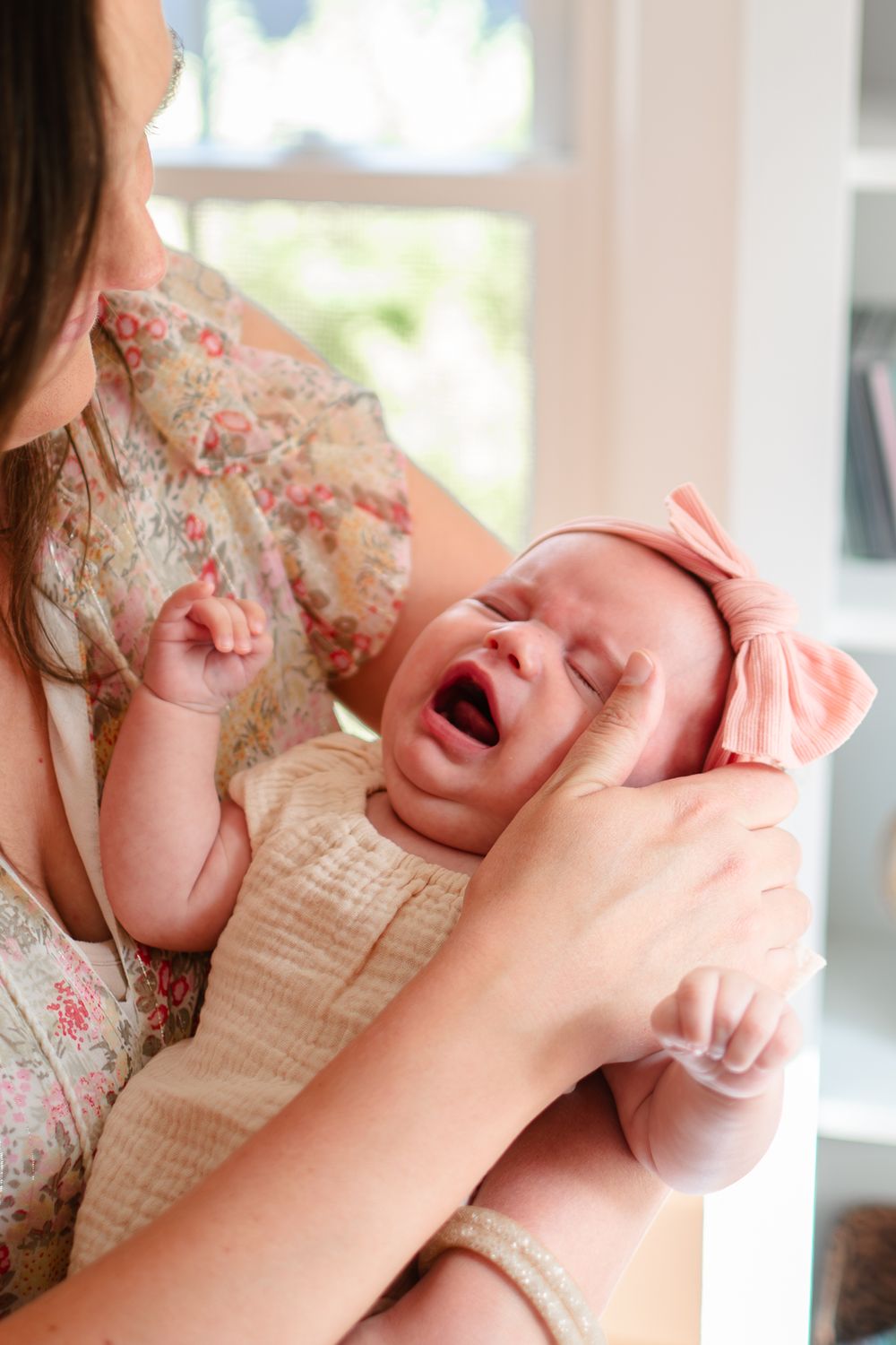 A crying newborn baby swaddled in a cream-colored blanket being comforted by gentle hands.