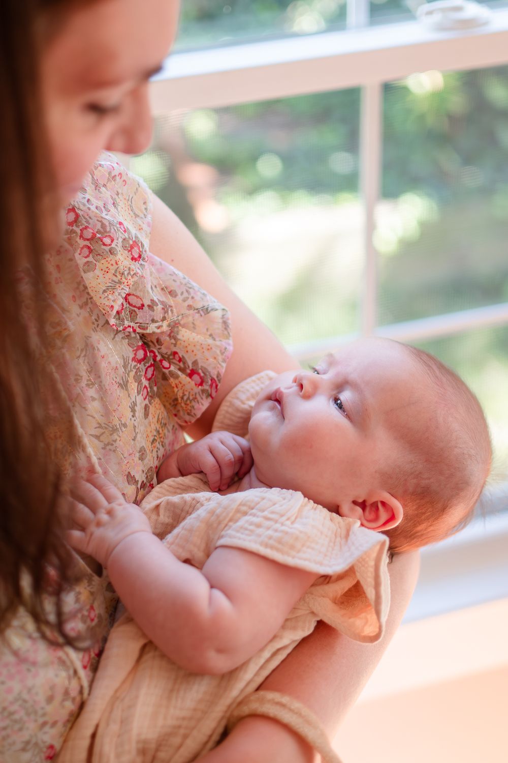 A baby in peach-colored clothing lies back in someone's arms near a sunlit window.