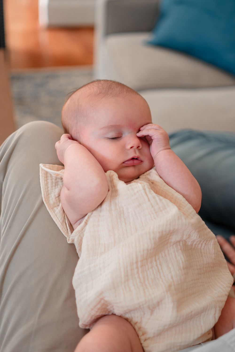 Sleeping baby in cream-colored muslin clothing rests peacefully on a gray cushion.