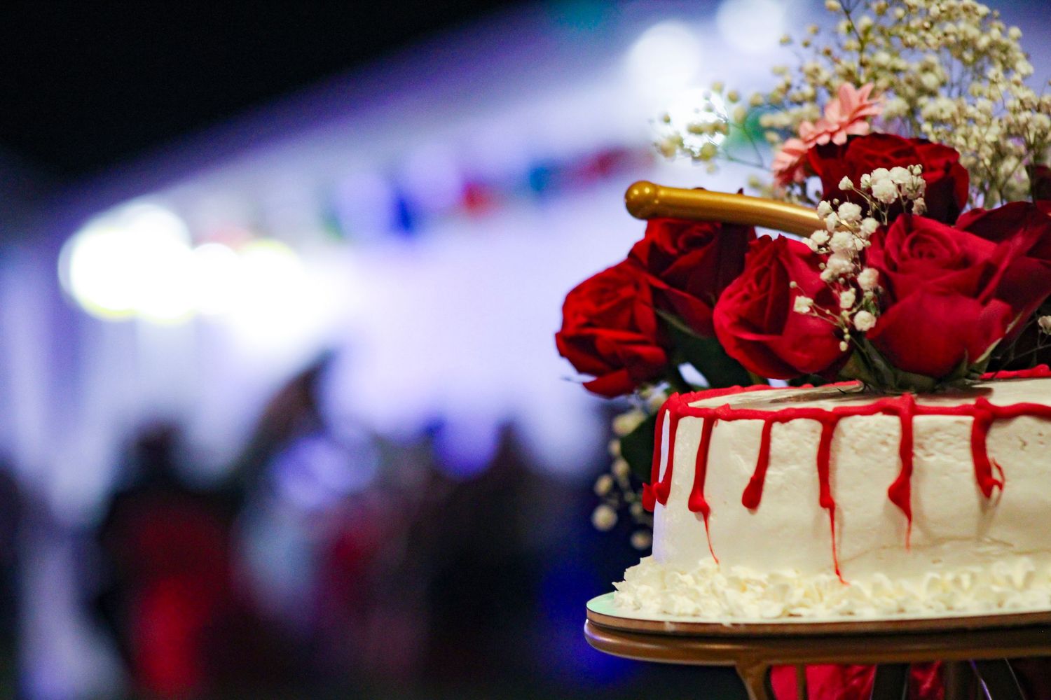 A red velvet drip cake decorated with fresh roses and white frosting sits on a dark display table.