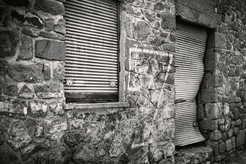 Two weathered windows with metal blinds set in an old stone wall photographed in black and white.