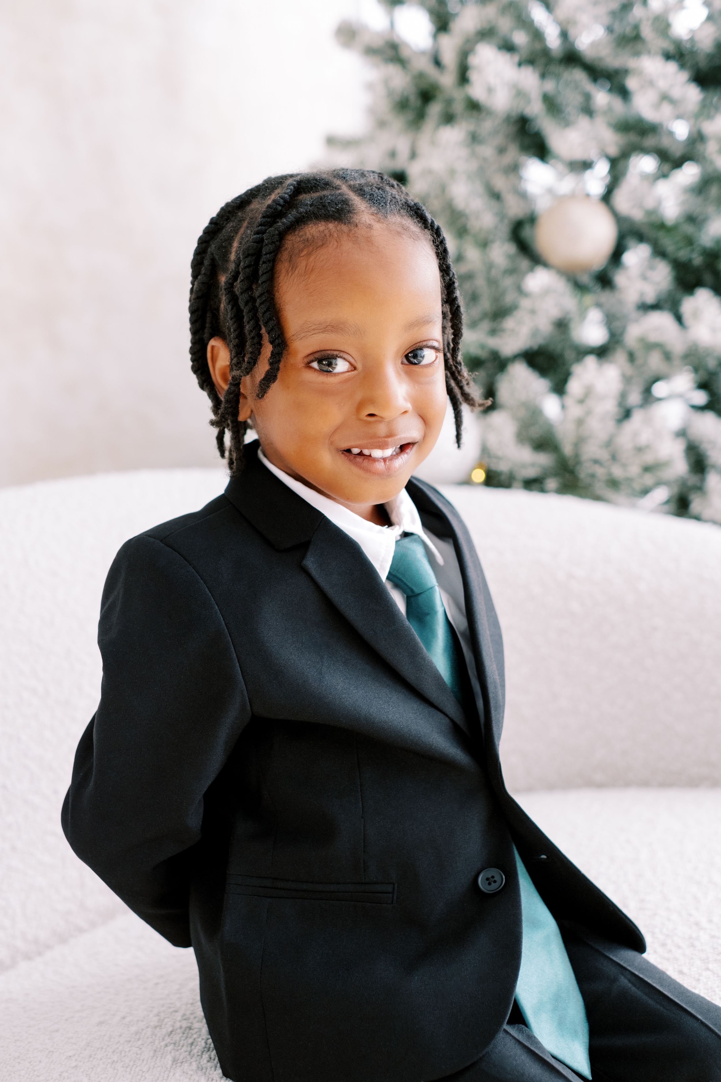 A young person in a black suit and turquoise tie poses with a bright smile against a light background.