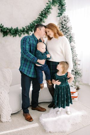 A family in coordinating green plaid and navy outfits poses together in a bright white studio with holiday decorations.