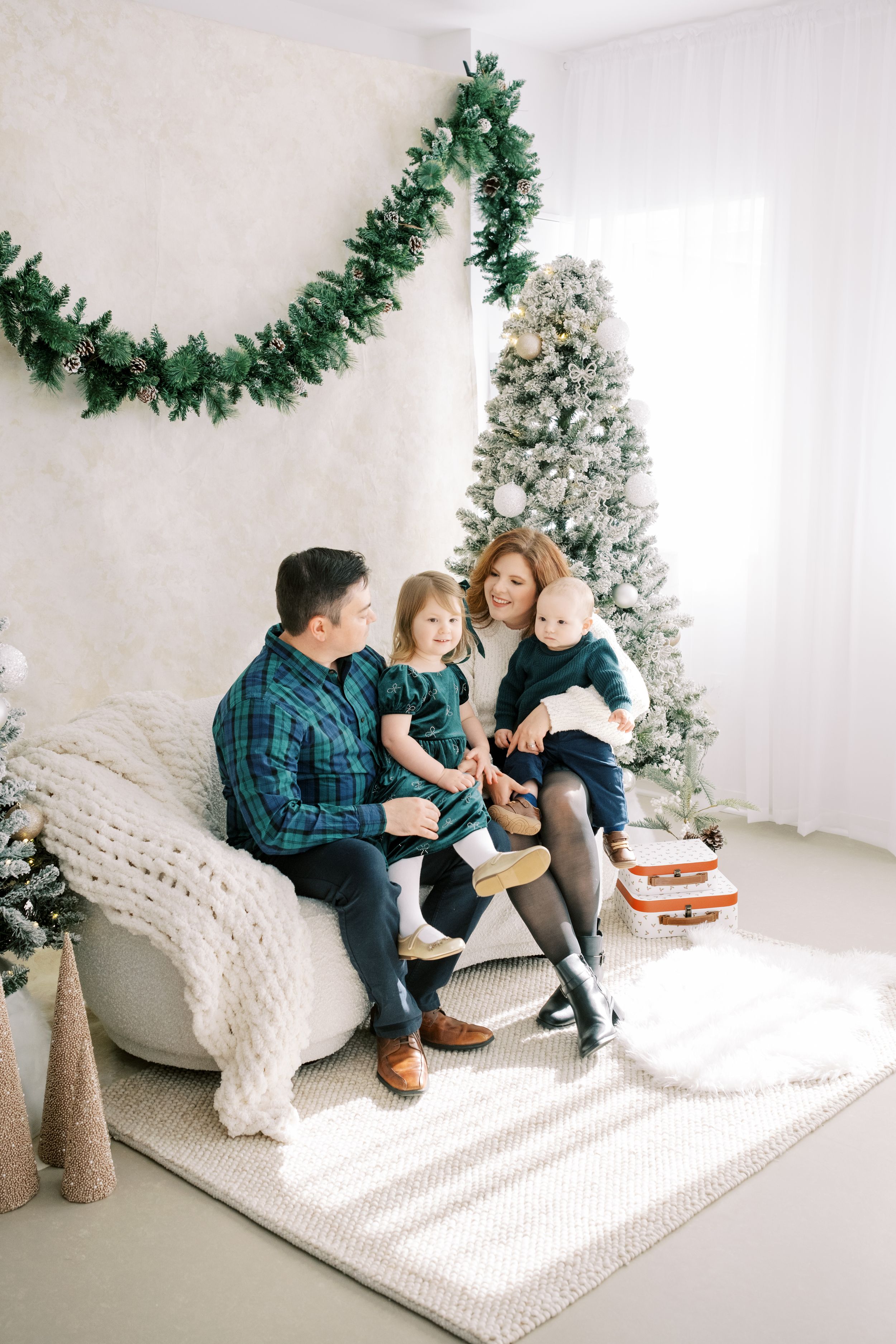 A family sits together on a white couch near a flocked Christmas tree with green garland and holiday decor.