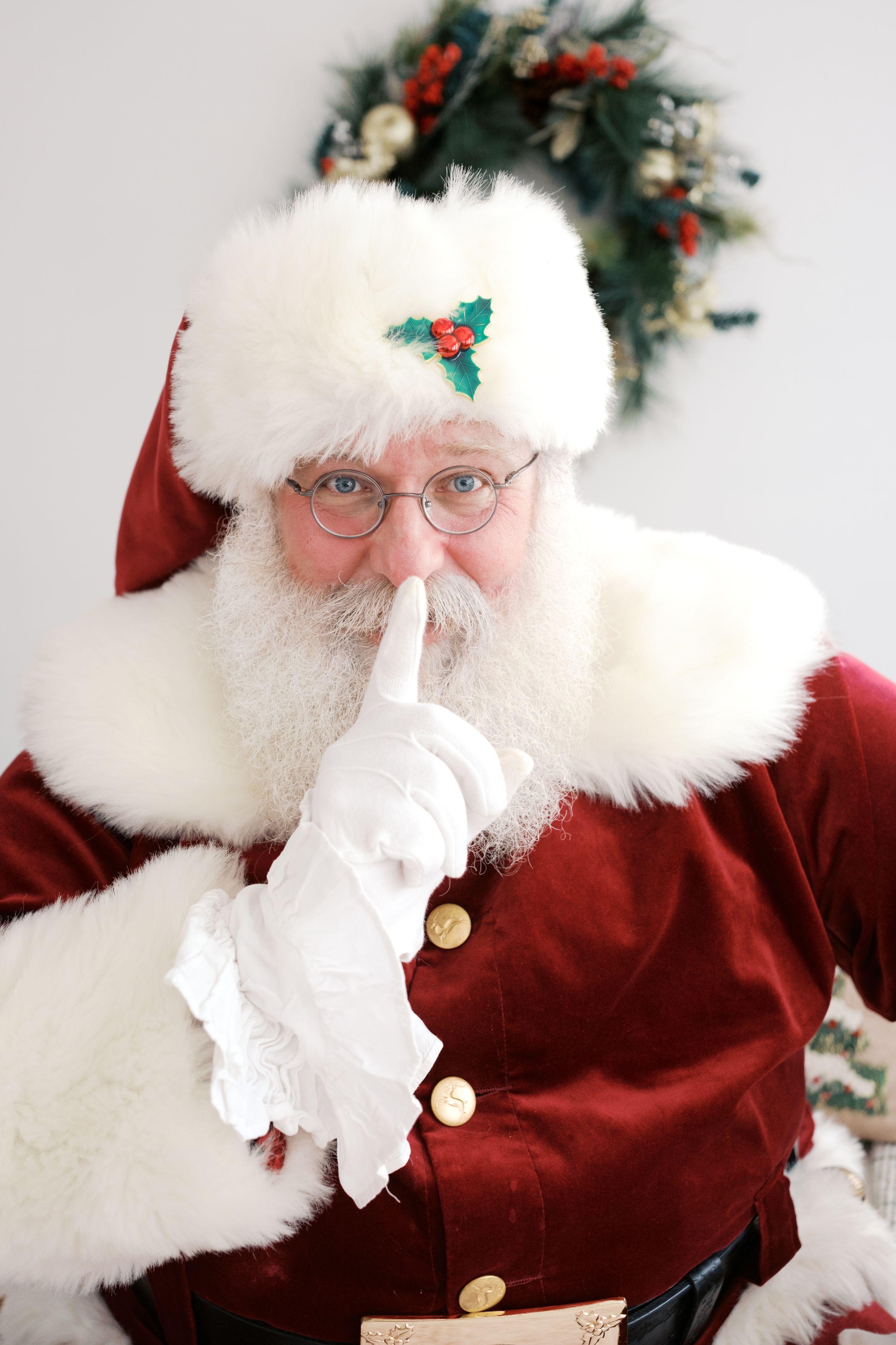 Santa Claus in red velvet suit with white gloves holds finger to lips in shushing gesture near Christmas wreath.