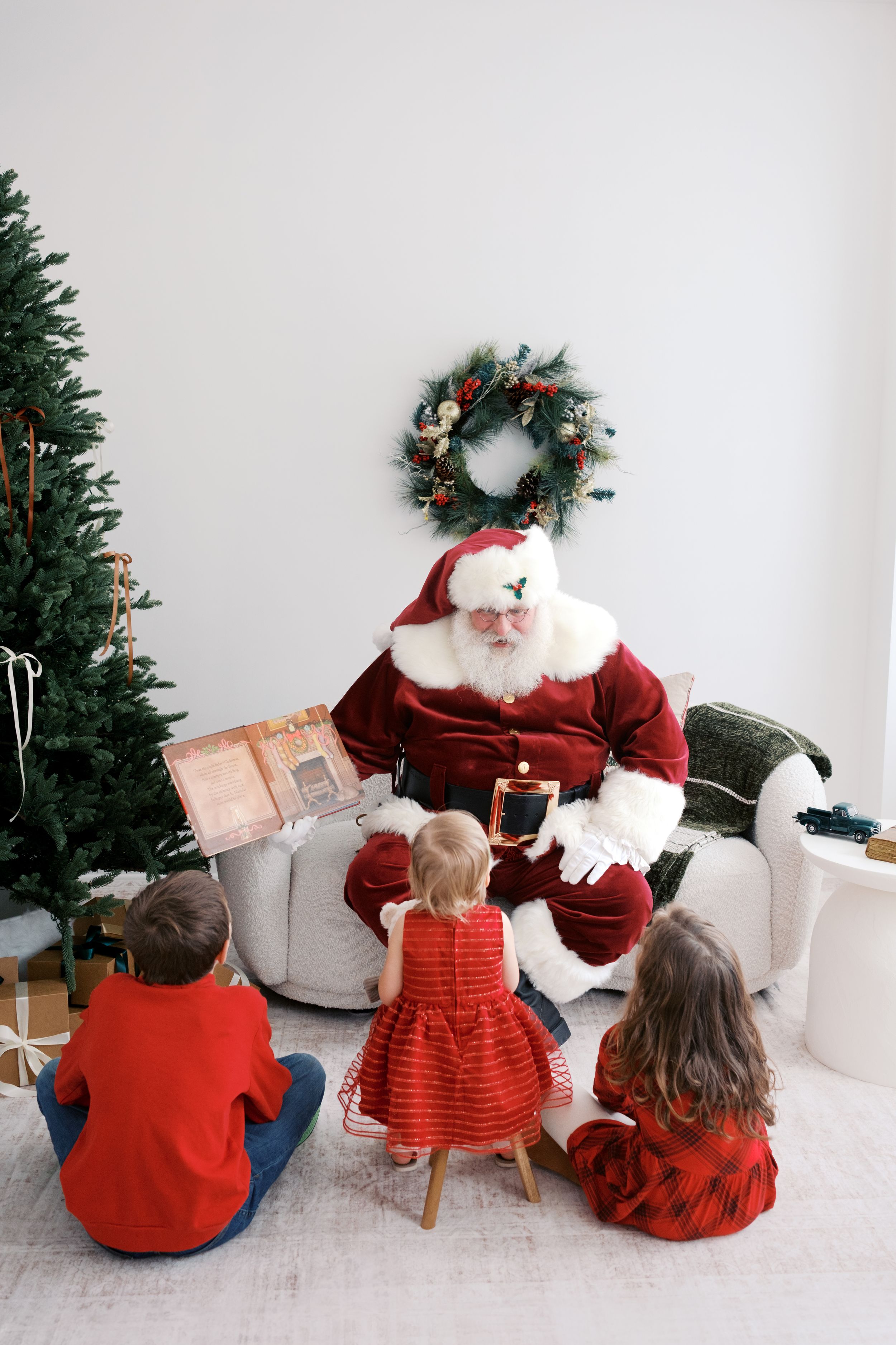 Santa Claus reads a book to three children seated on the floor near a decorated Christmas tree and holiday wreath.