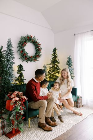 Family sits together on couch surrounded by Christmas trees, wreath and holiday decorations in bright white room.