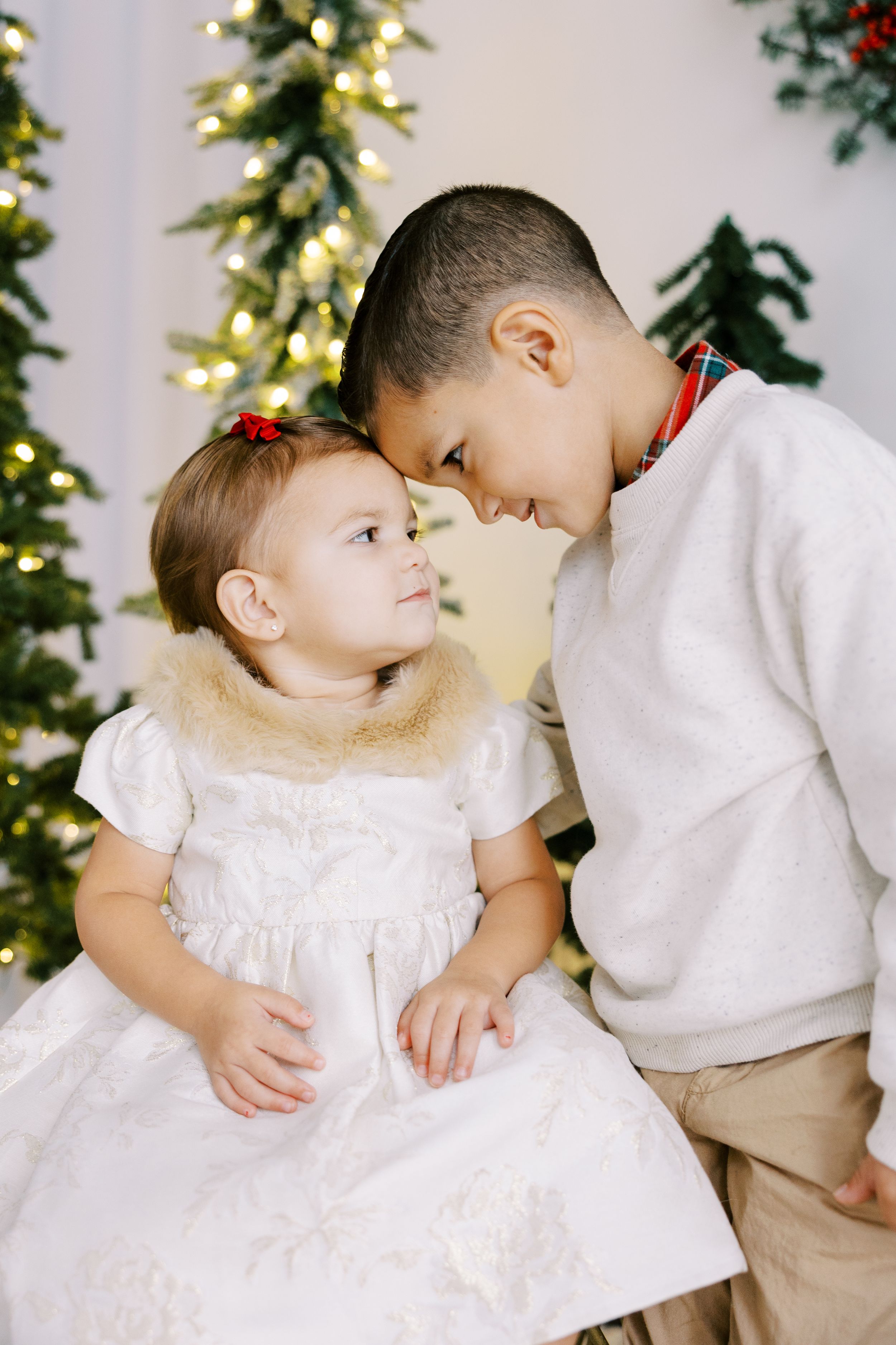 Two young children in holiday attire share a sweet moment in front of a decorated Christmas tree with twinkling lights.