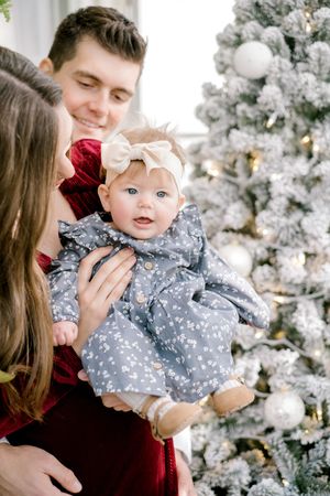 Family poses for a holiday portrait in front of a white flocked Christmas tree with a baby wearing a gray floral dress.