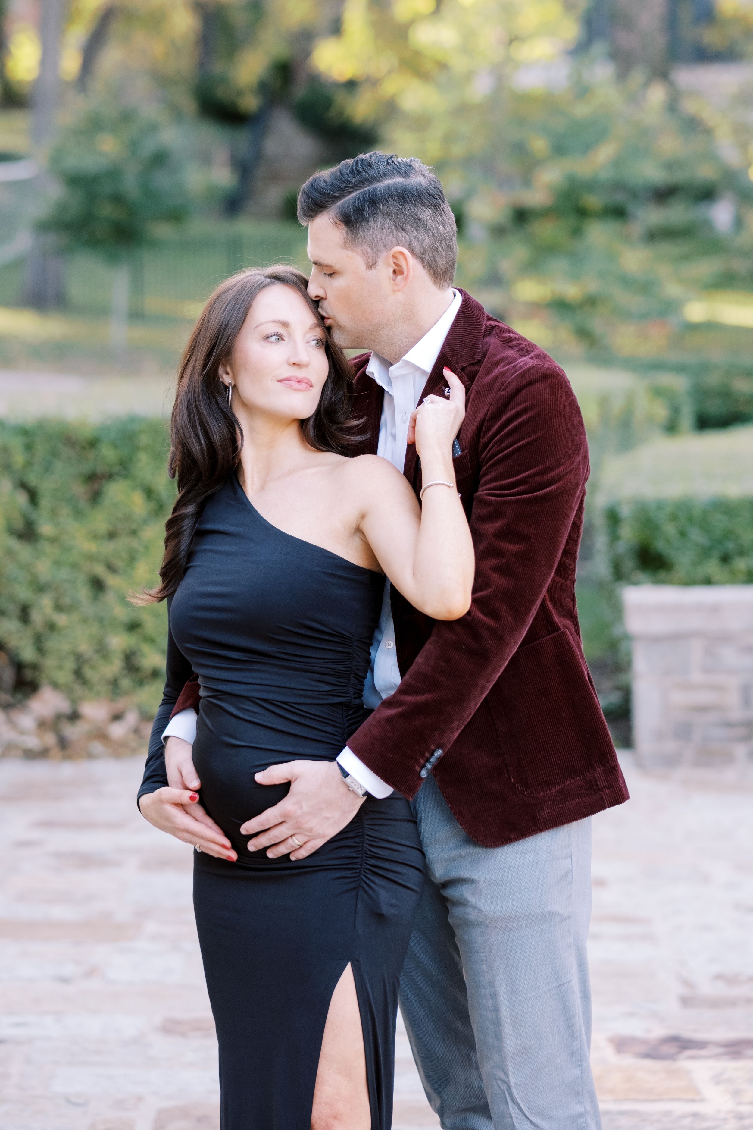 A romantic couple embraces outdoors, with one wearing a burgundy velvet jacket and the other in an elegant black dress.