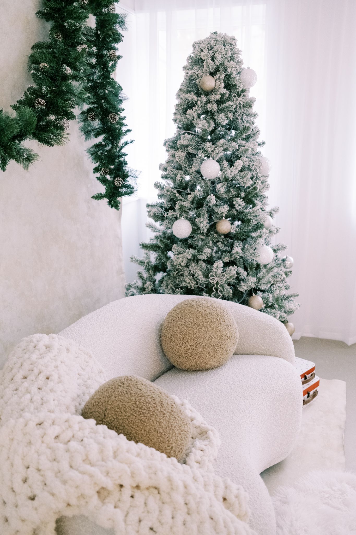 A white knitted blanket with a tan pillow sits beneath snow-dusted evergreen trees in a minimalist winter scene.