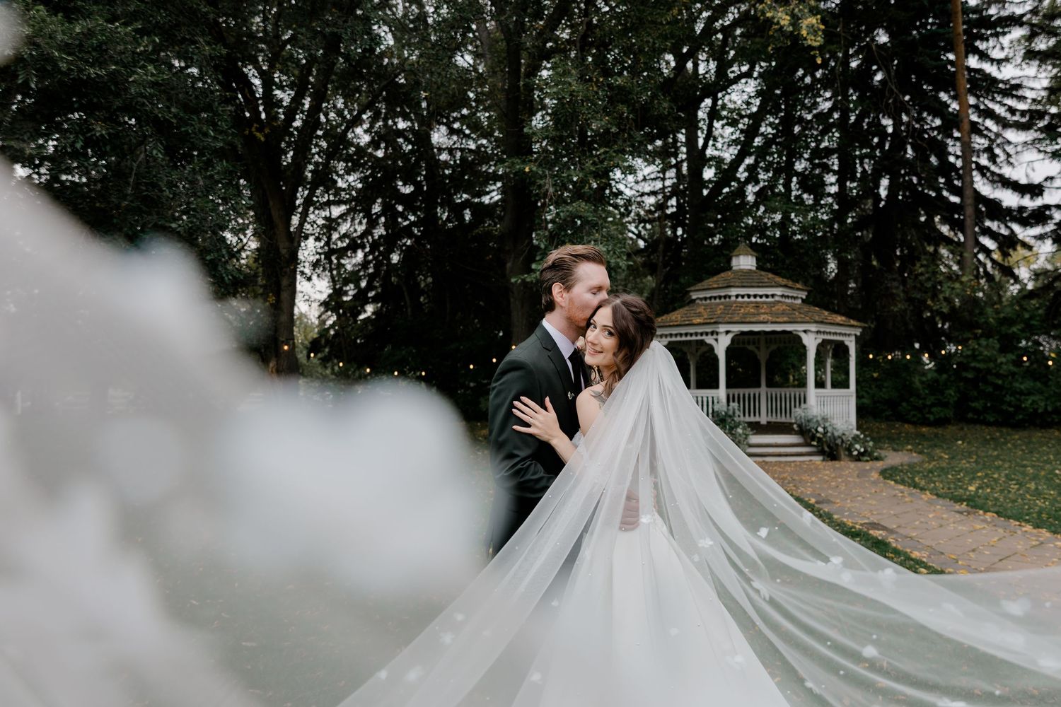 A couple shares an intimate moment in front of a white gazebo while a long flowing veil trails behind the bride.