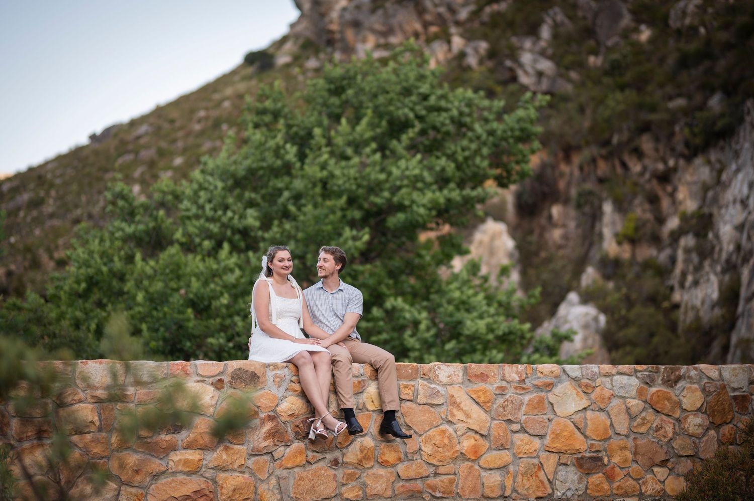 A couple sits together on a stone wall with a scenic mountain landscape in the background.