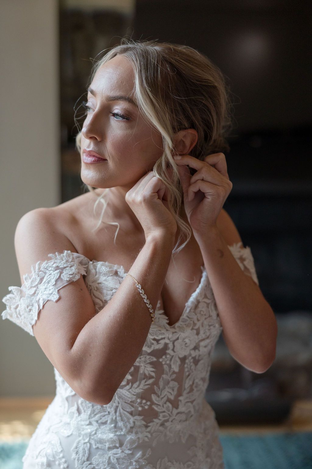A bride in a delicate lace off-shoulder wedding dress adjusts her earring in soft natural light.
