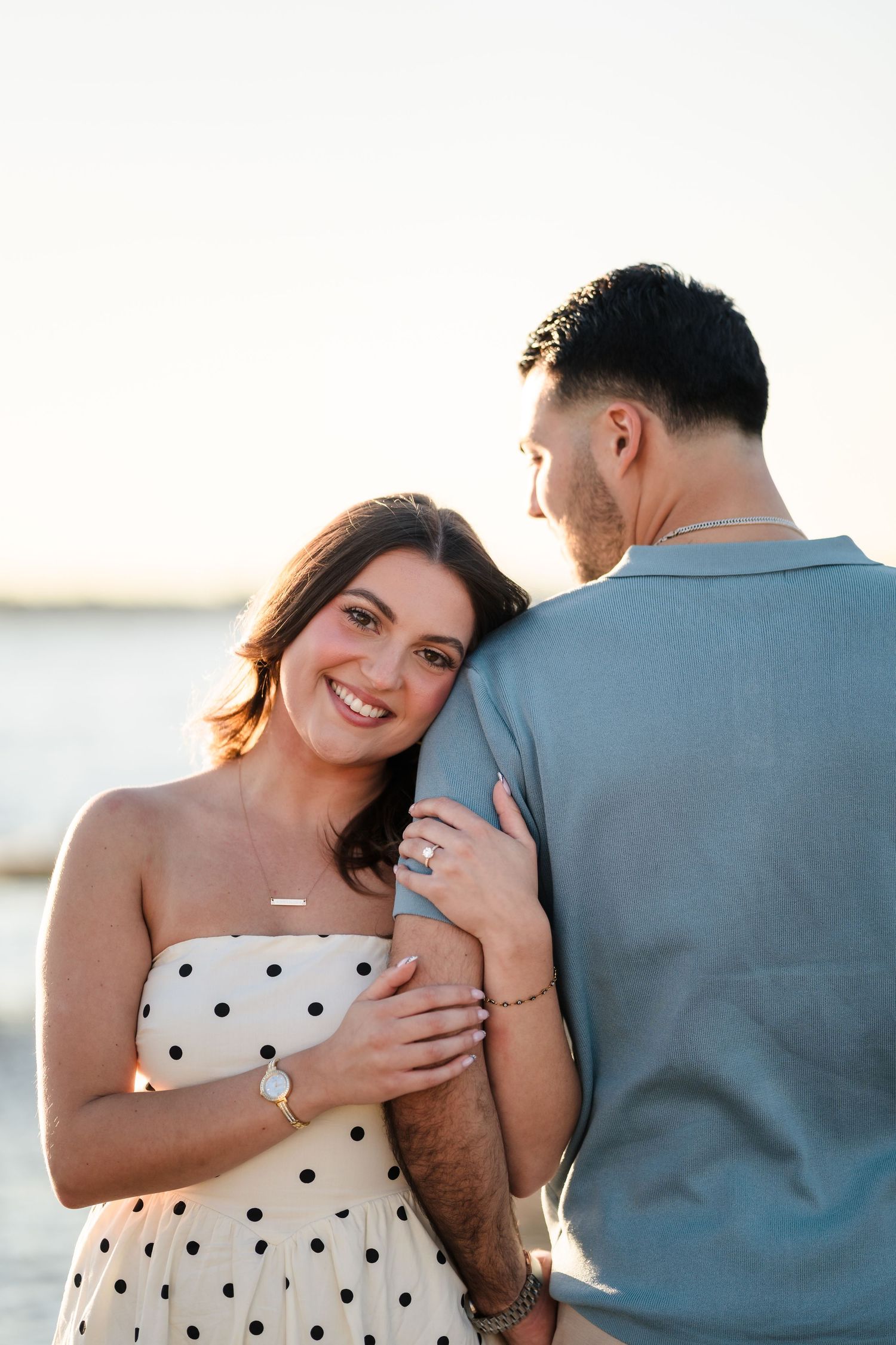 A couple embraces by the water, with one wearing a polka dot dress.