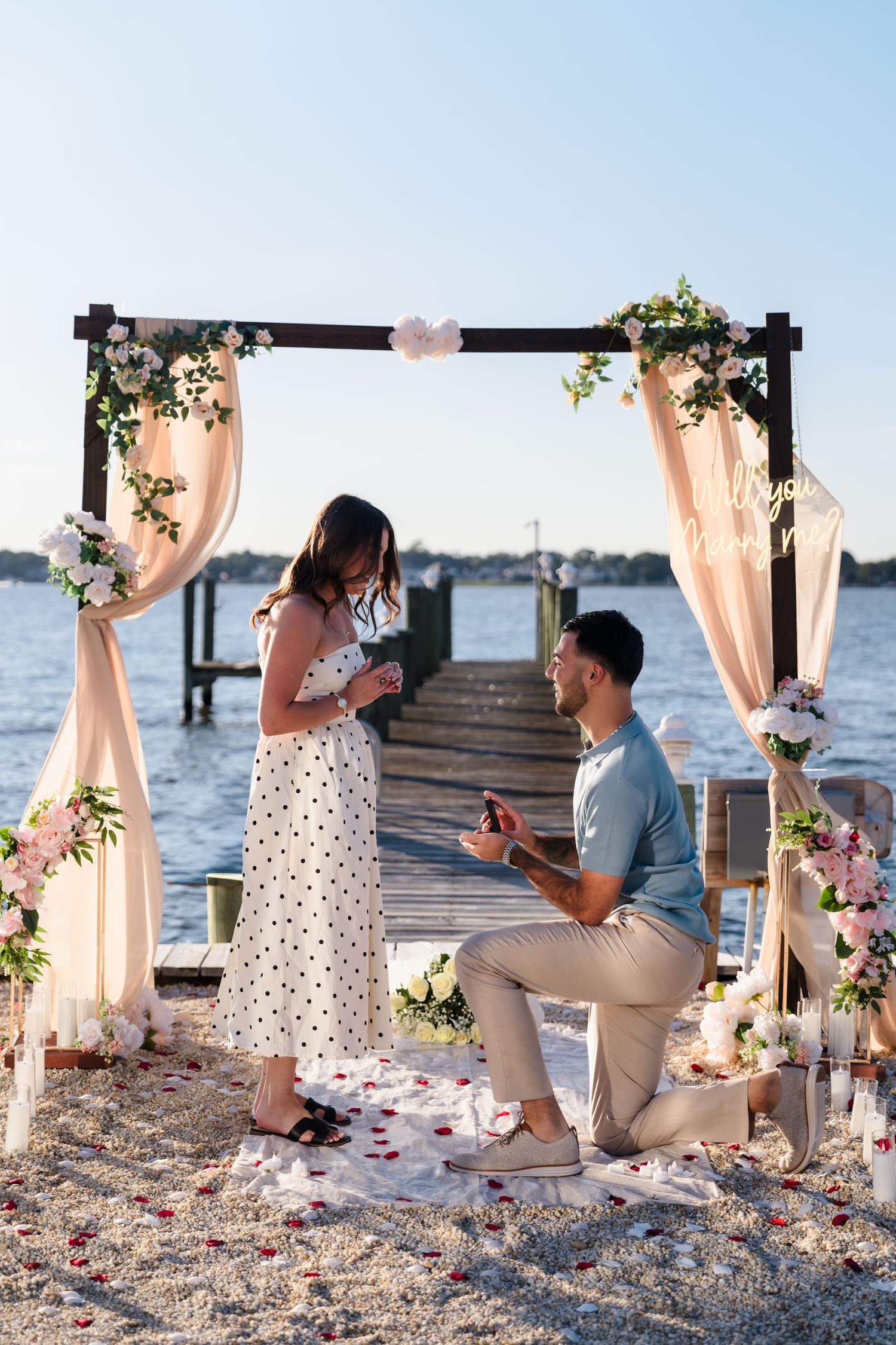 A romantic beach proposal setup with a decorated wooden arch and rose petals scattered on the sand at sunset.