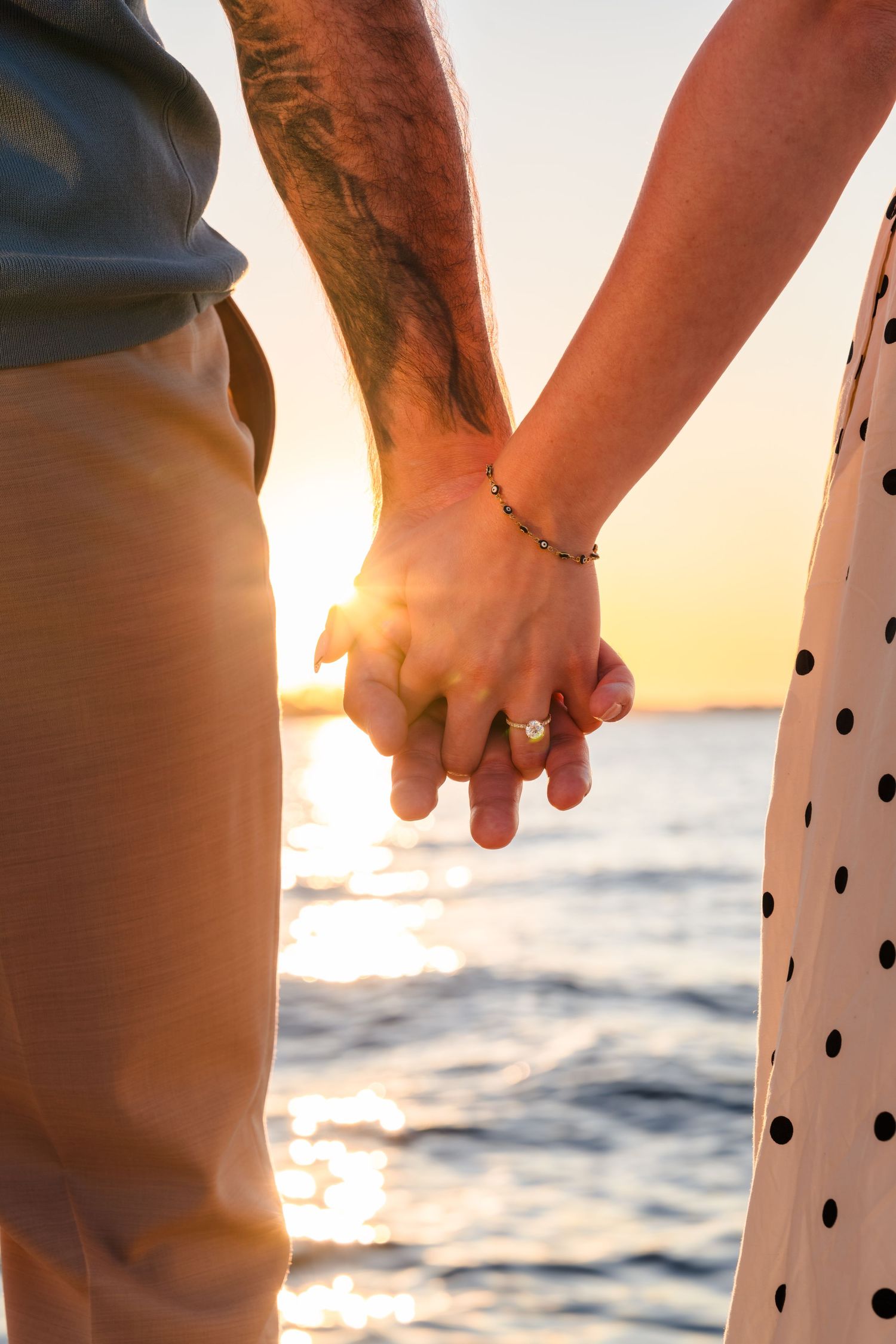 Close-up of hands holding while wearing polka dot dress against sunset ocean backdrop.
