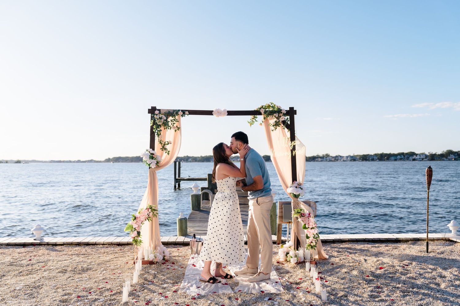 Beachside proposal scene with draped fabric and flowers on a wooden arch overlooking the water.