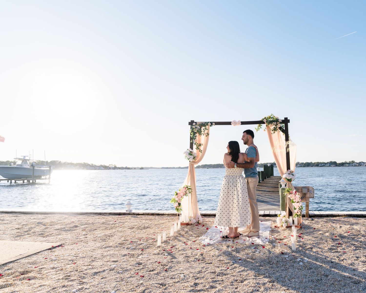 Scenic waterfront proposal setting with white curtains and floral decorations at golden hour.