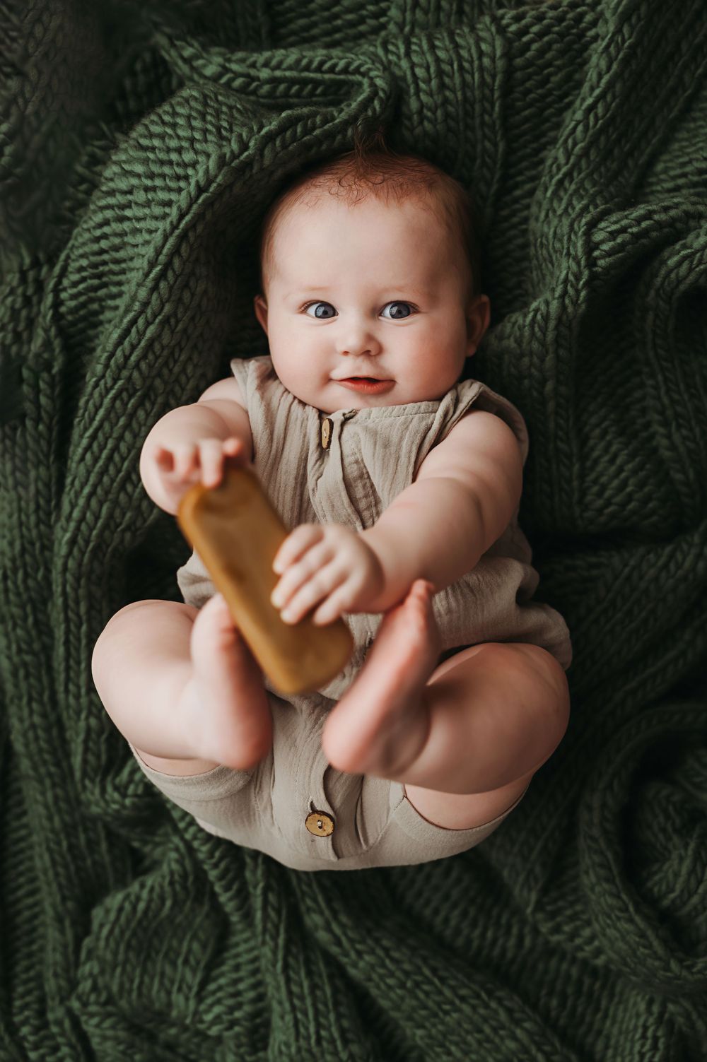 Baby in beige romper lies on dark green knitted blanket while holding wooden toy.