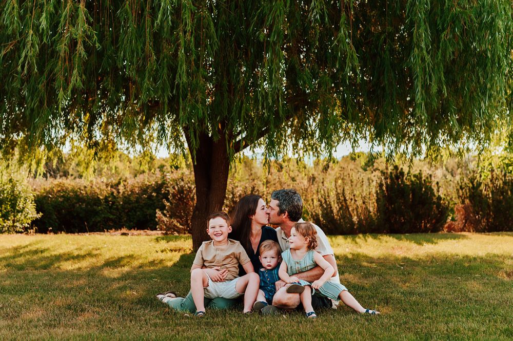 A family sits together on grass under a willow tree during a golden hour photo session in a park.