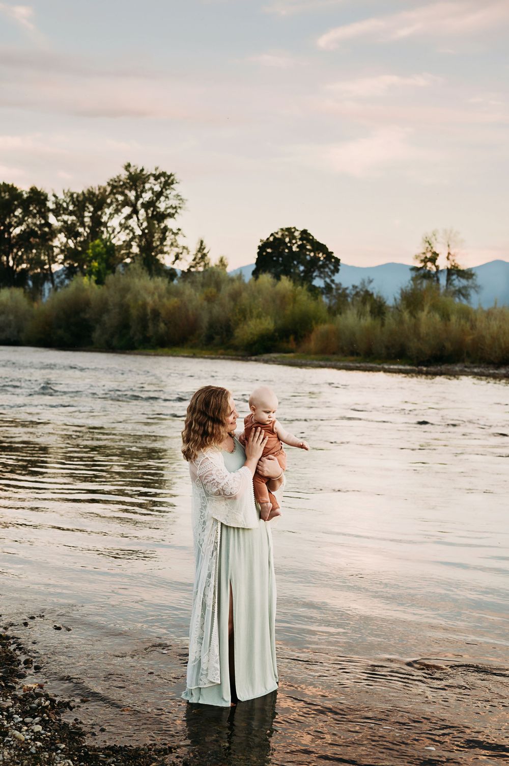 Two figures in light clothing embrace by a river at sunset with mountains and trees in the background.