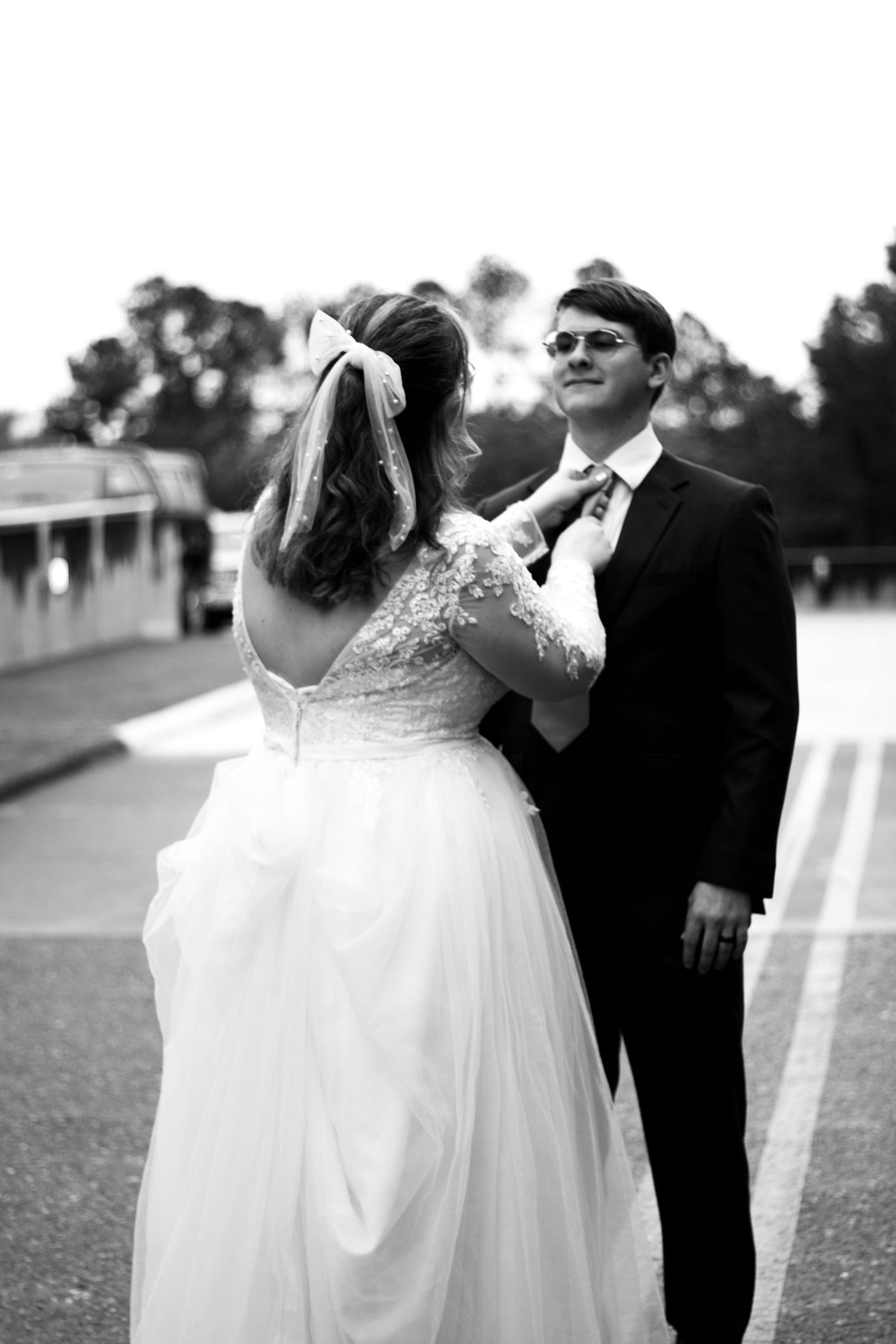 Black and white photo of a bride in a flowing white dress and groom in sunglasses sharing a romantic moment outdoors.