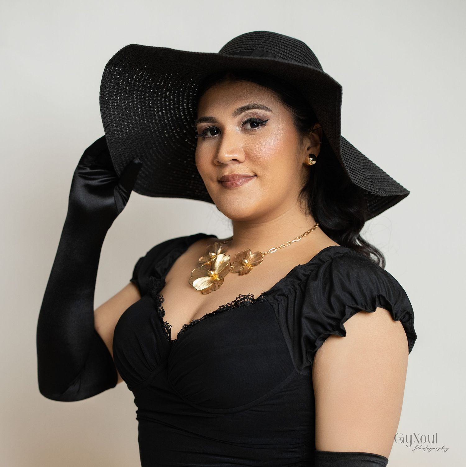Elegant birthday portrait in black dress with wide-brimmed hat and long gloves against neutral background.