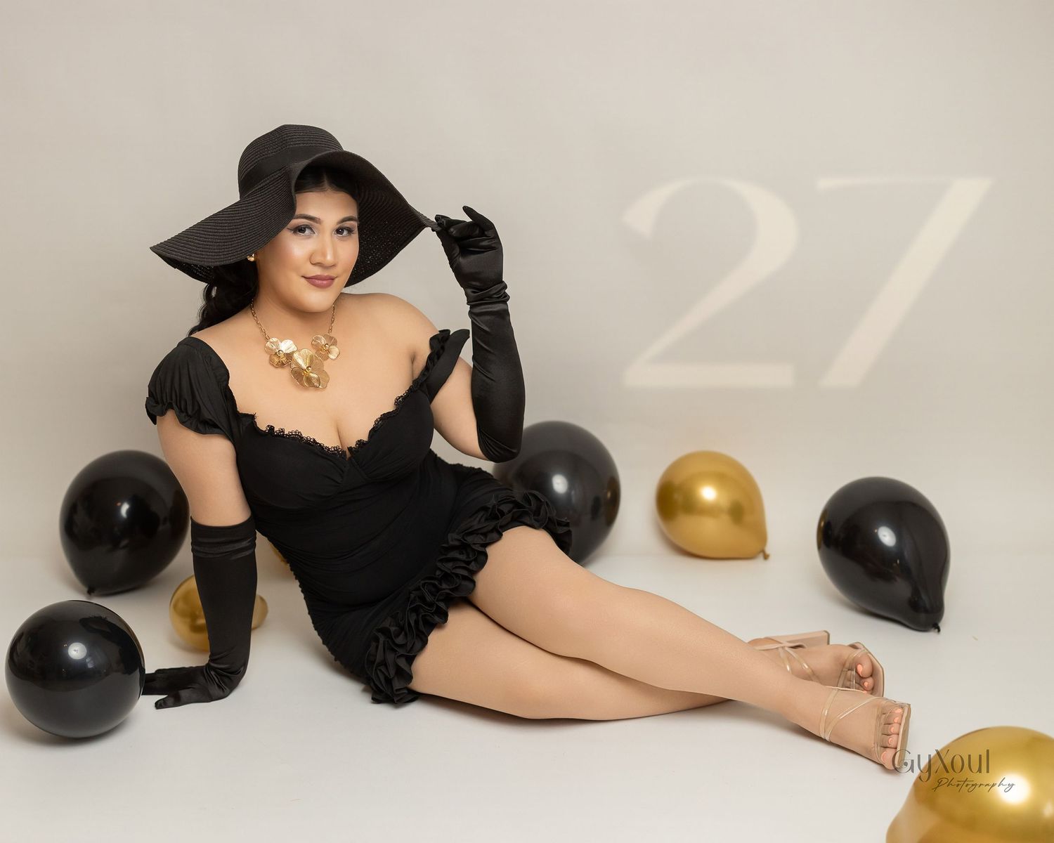 Glamour birthday photo with black dress, hat and gloves posed with gold and black balloons.
