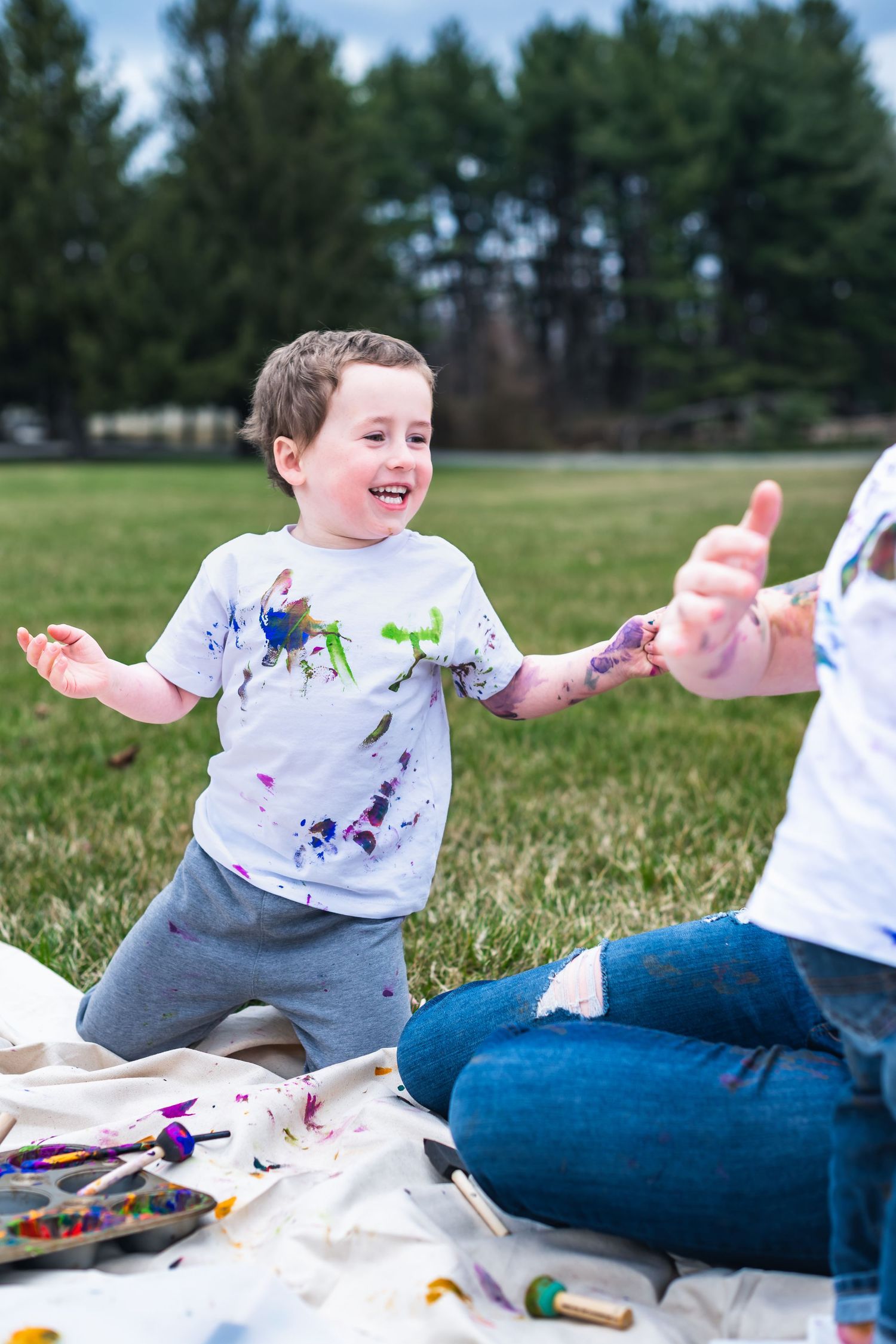 Young child playing with paint outdoors on a blanket, wearing a messy white t-shirt while laughing and gesturing.