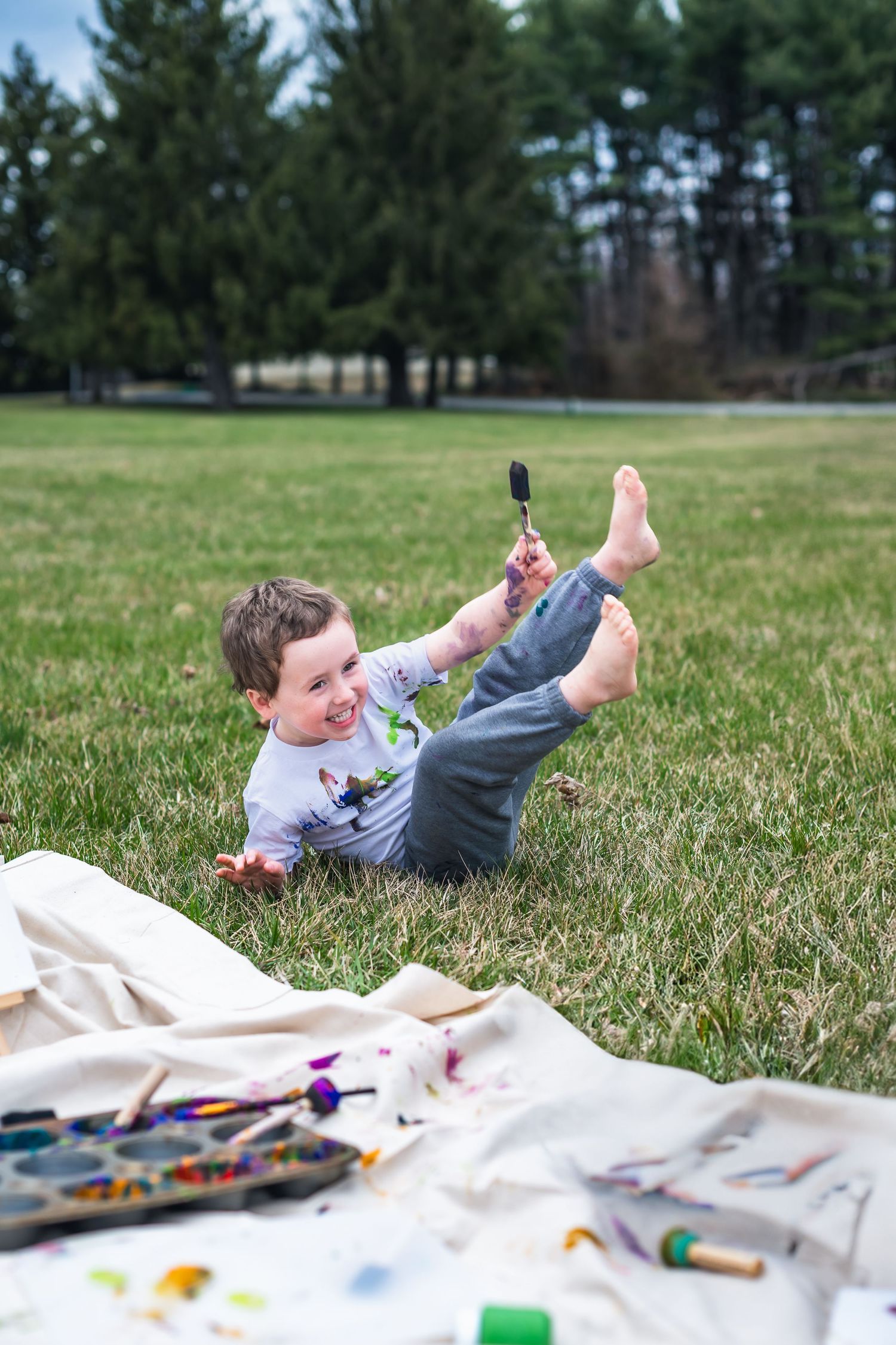 Child lies on grass near art supplies spread out on blanket during outdoor creative time on sunny day.