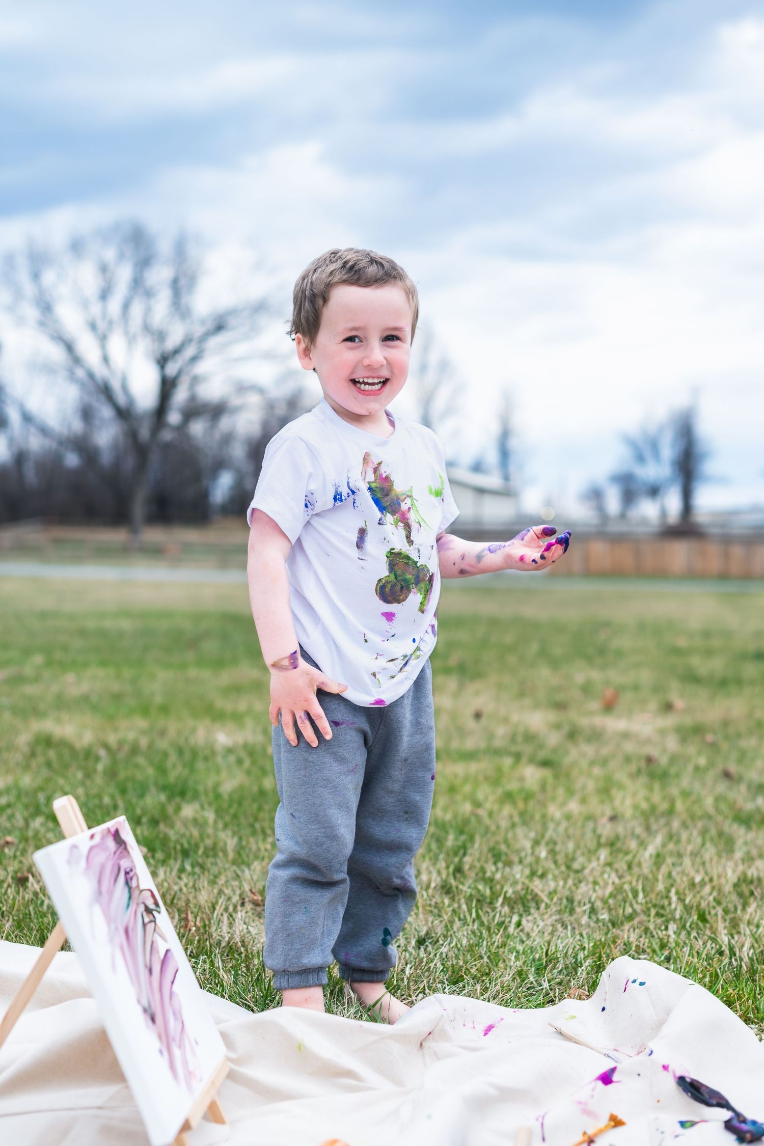 A young child in a colorful paint-splattered shirt stands on grass near an outdoor art project on a sunny day.