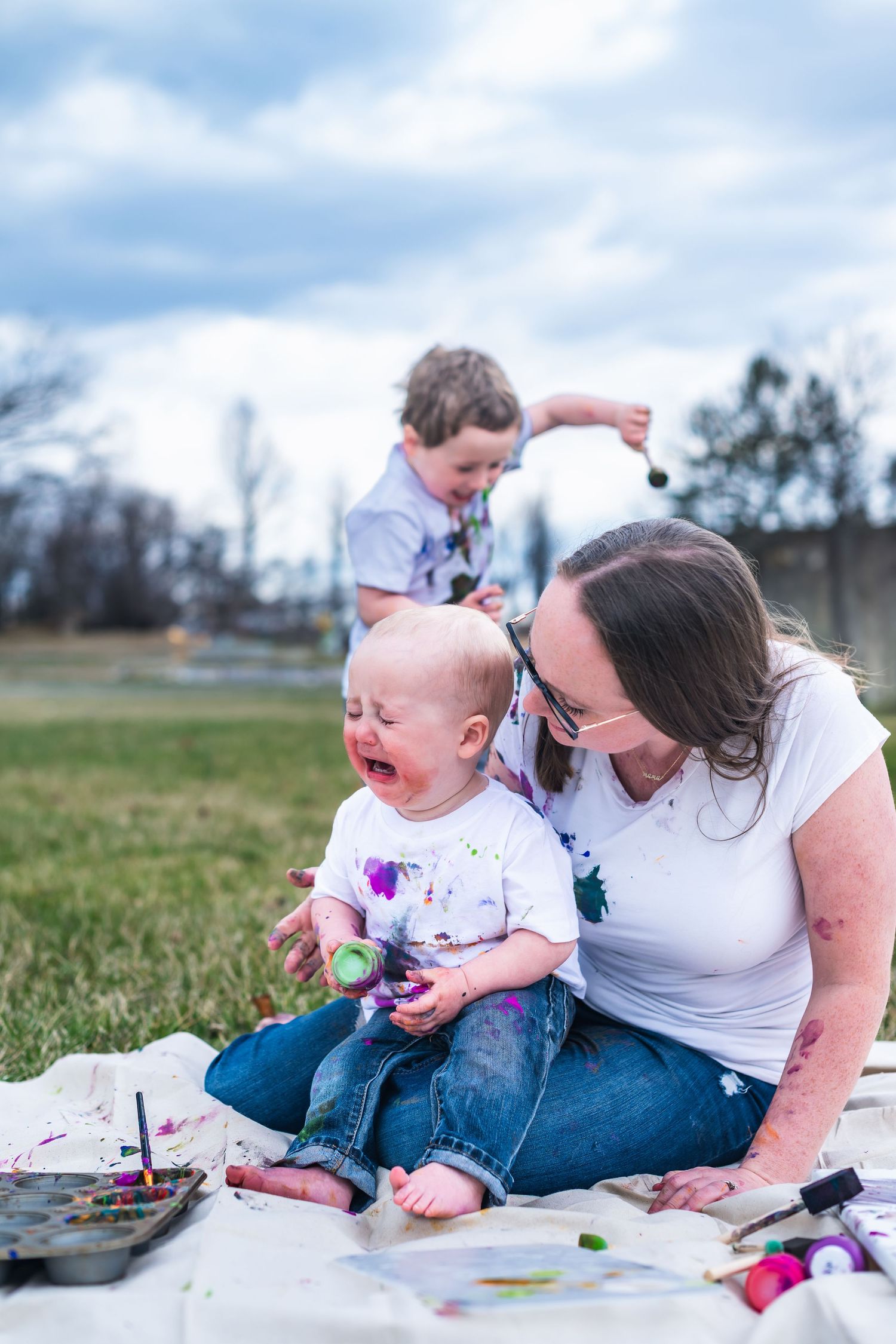 A joyful outdoor art session with colorful paints spread on a blanket in a grassy field.