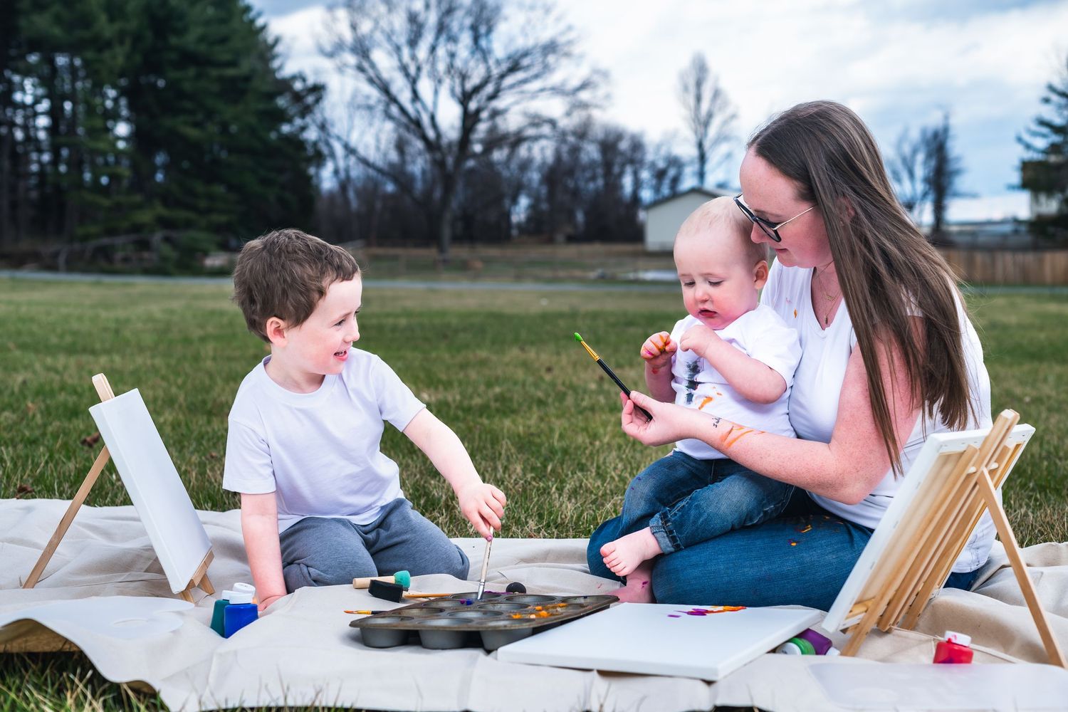 People enjoying outdoor art time on a blanket with painting supplies and toys spread out in a grassy field.