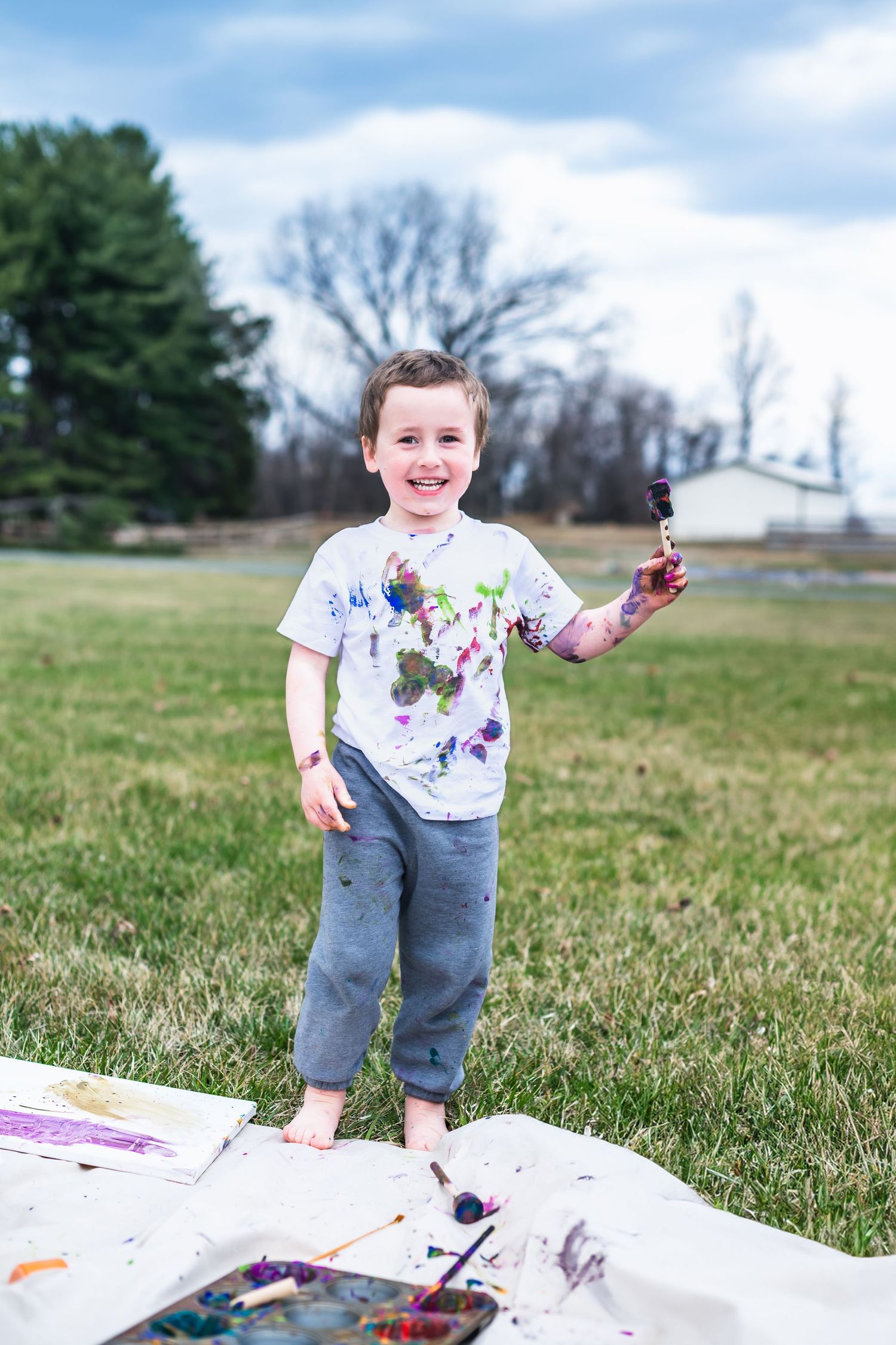 Young artist outdoors holding a paintbrush with colorful paint stains on white shirt standing on a blanket in grass field.