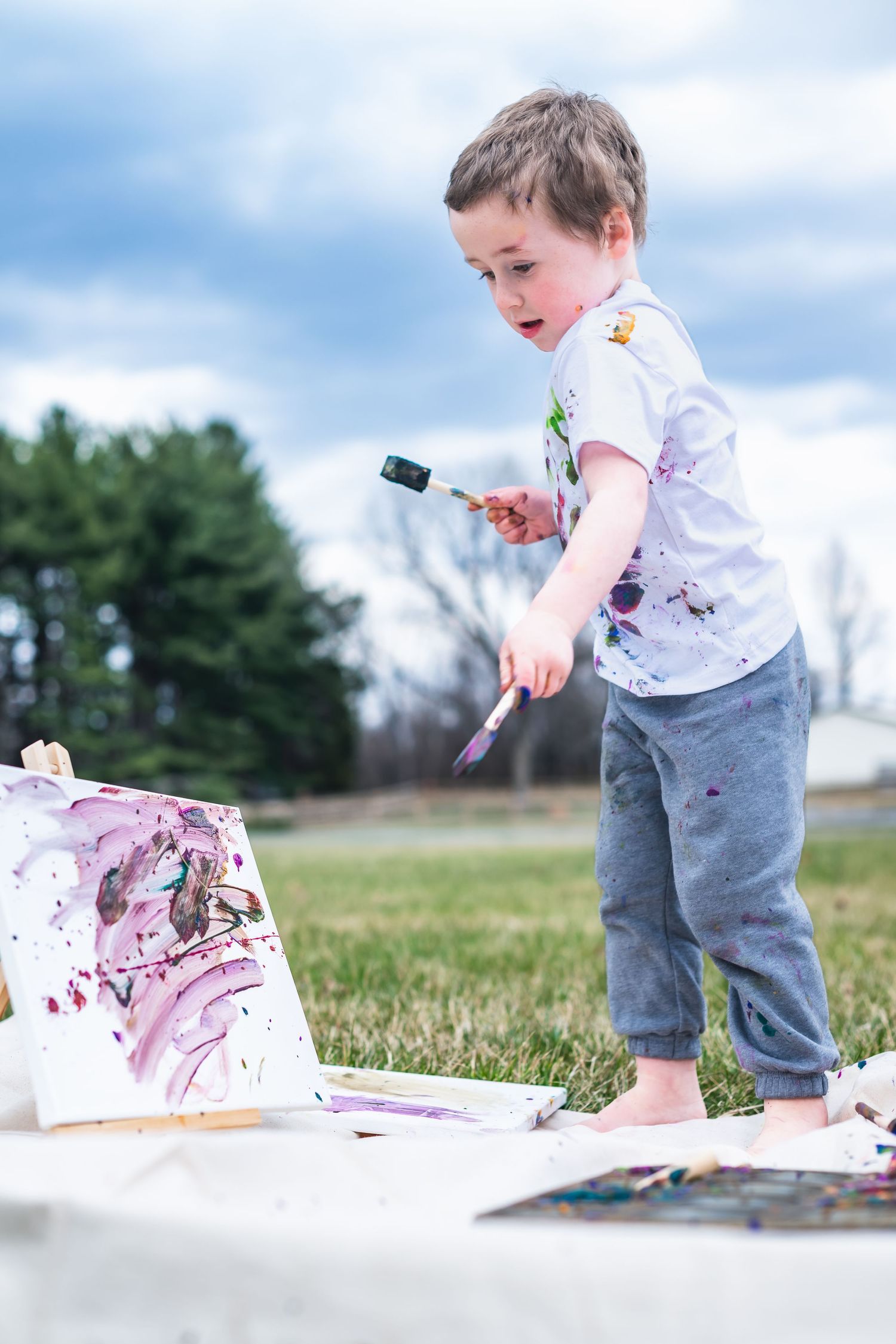 A young child in paint-splattered clothes creates outdoor artwork on a blanket during a sunny day.