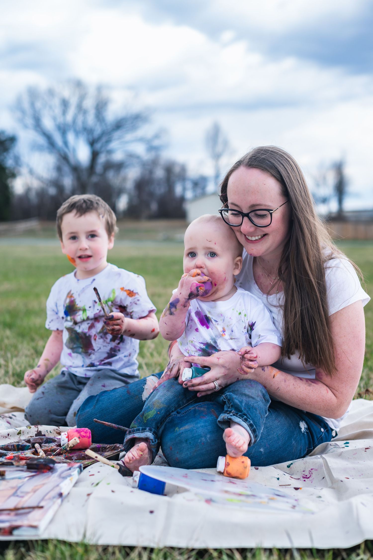 Family enjoying messy outdoor art time on a blanket in a grassy field on a partly cloudy day.