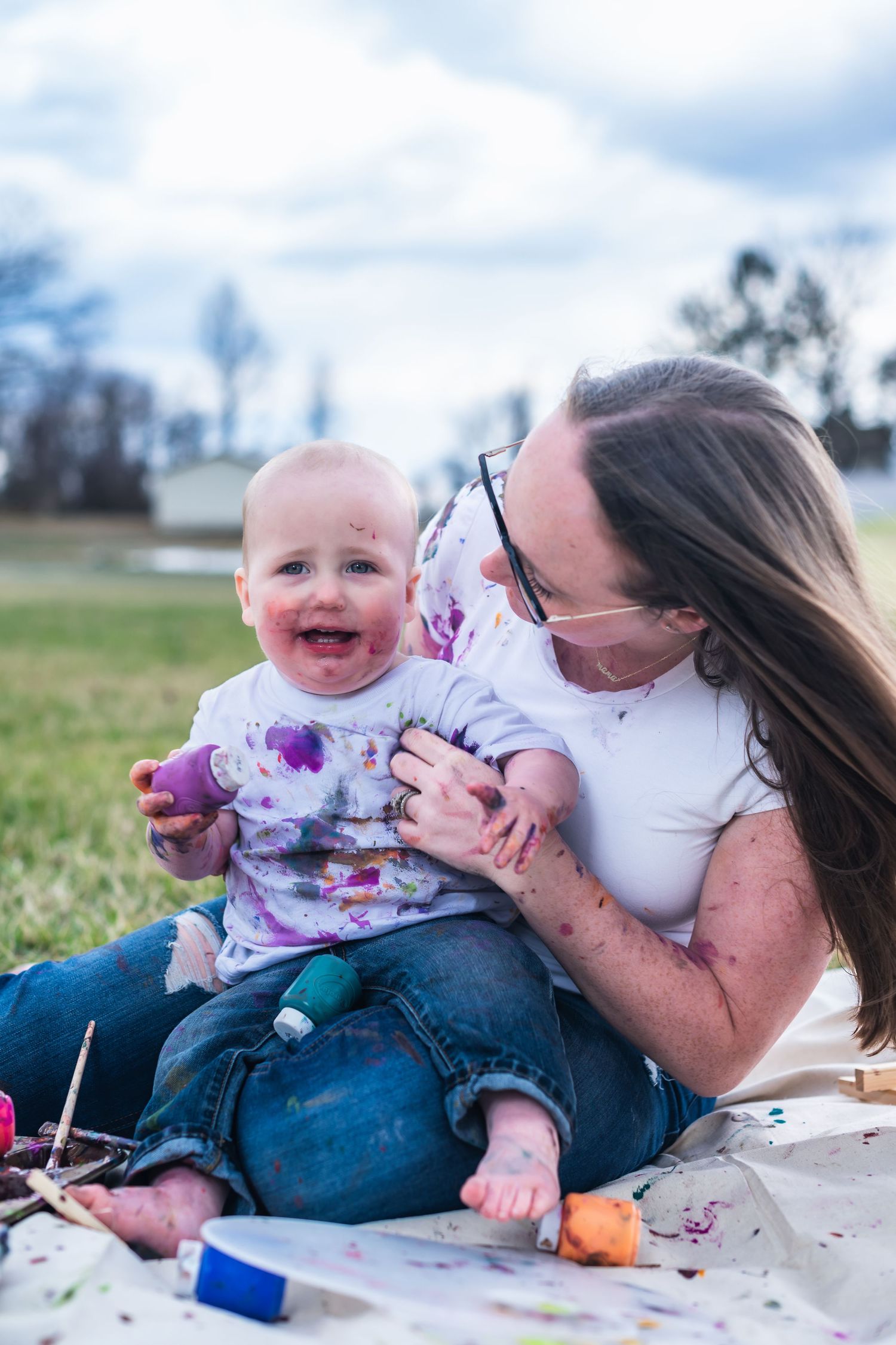 Parent and child share a joyful moment while sitting on a blanket outdoors with art supplies scattered around.
