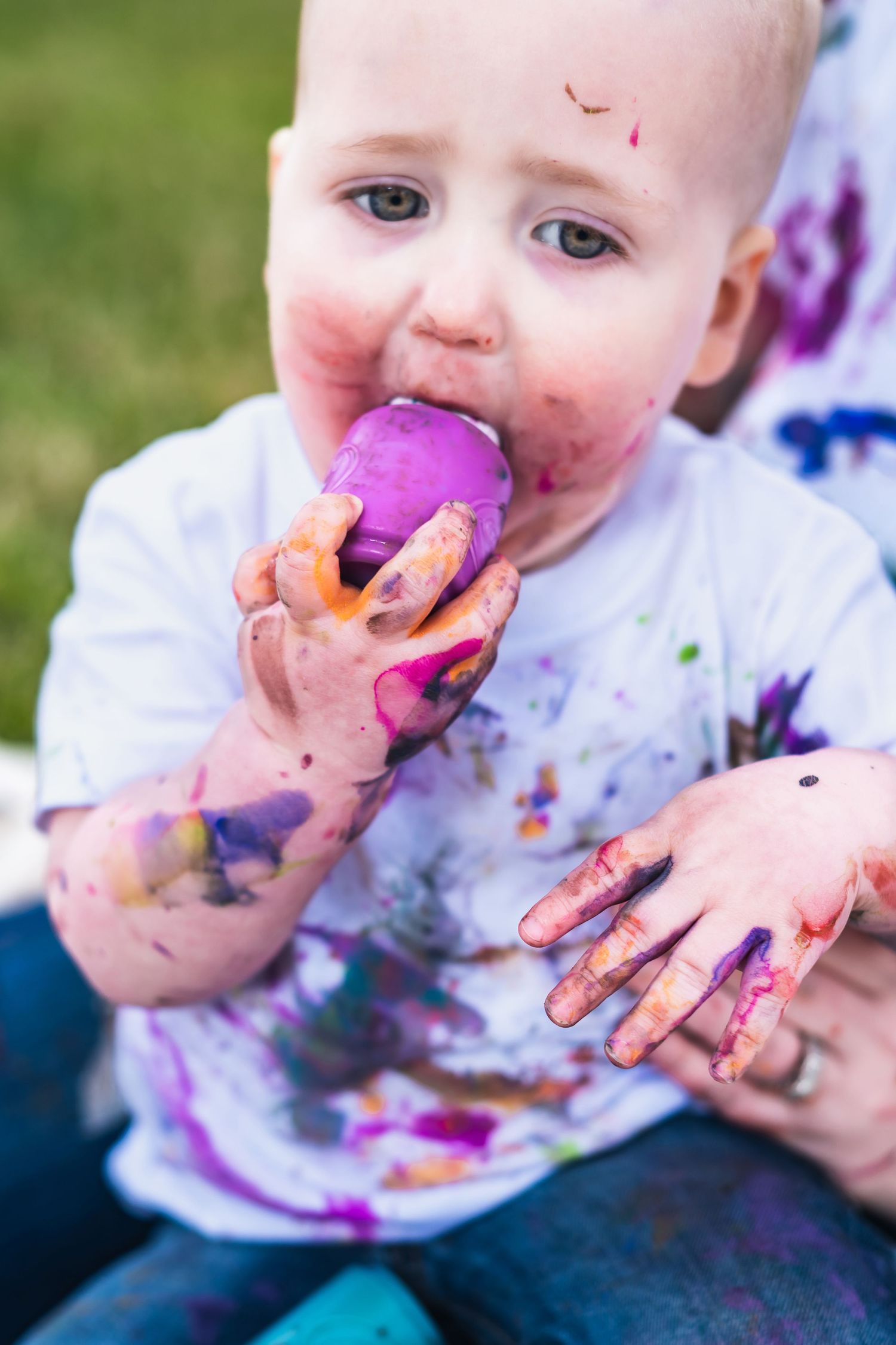 Messy toddler in white shirt covered in colorful paint holds purple snack while sitting outside on grass.