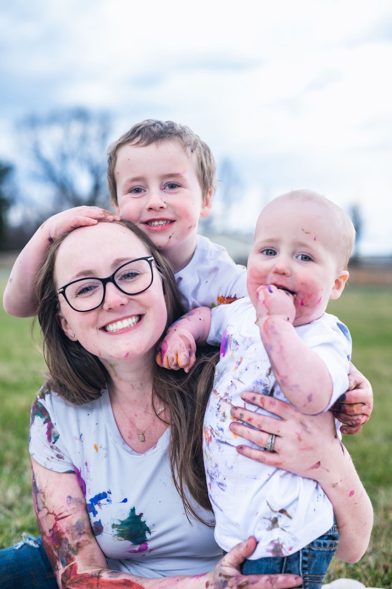 A cheerful family sits together outdoors on a sunny day wearing matching paint-splattered white t-shirts.