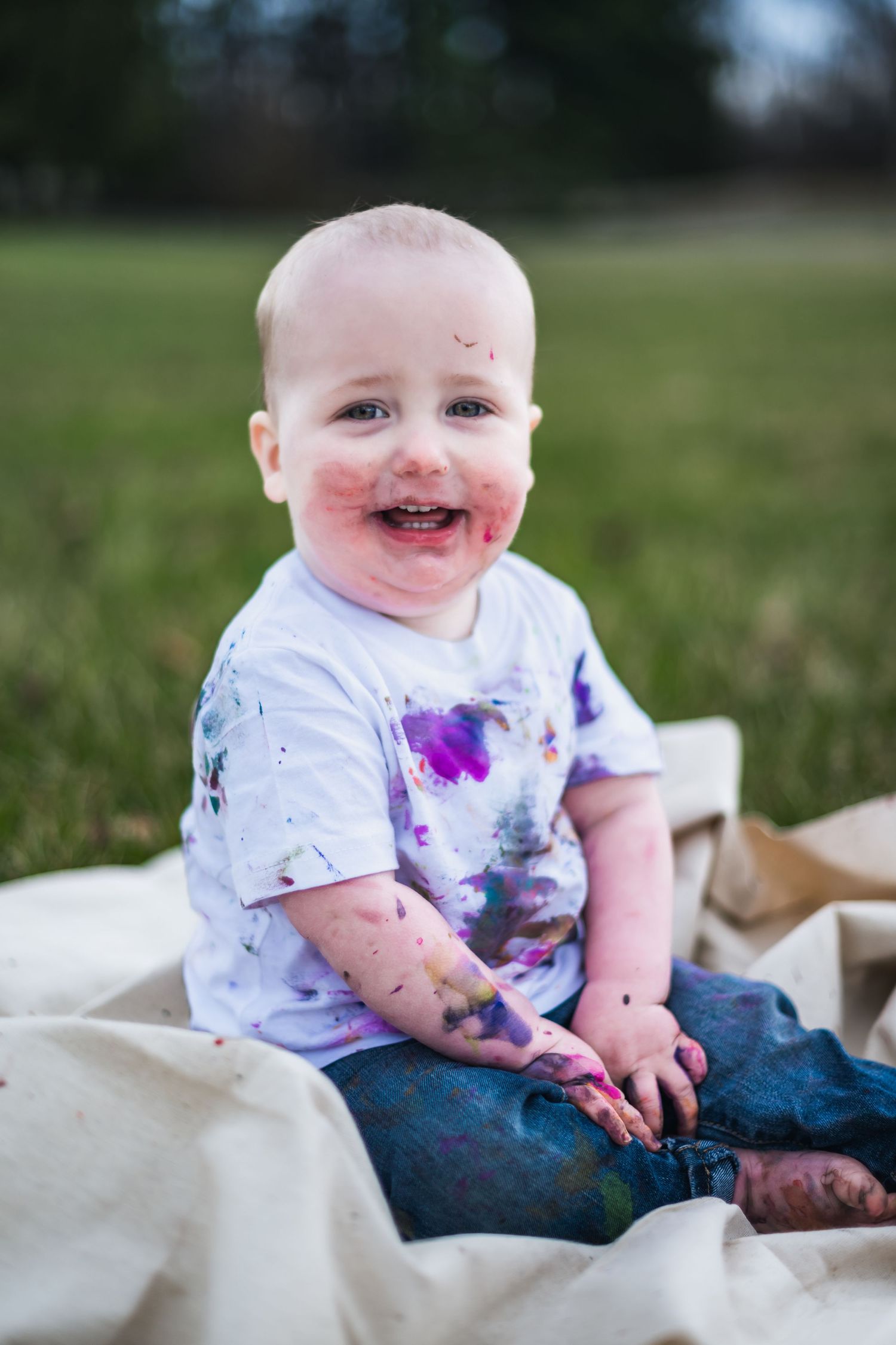 Happy baby on white blanket with purple paint stains on clothes and face sits outdoors in green grass.