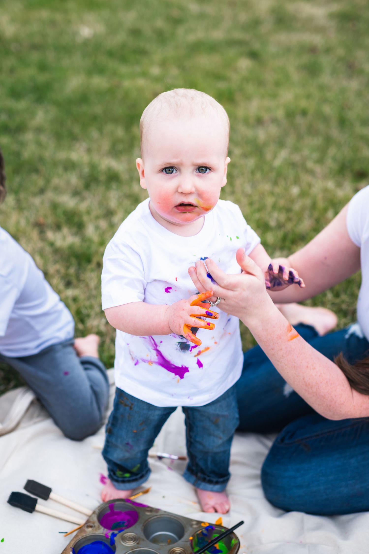 A toddler in a white t-shirt and jeans stands on a blanket with paint supplies during an outdoor art activity.