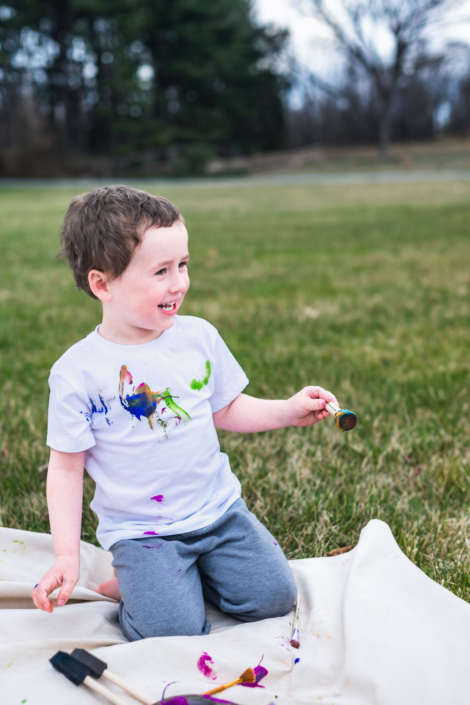 Young child wearing a dinosaur t-shirt sits on a blanket outdoors playing with colorful objects on a grassy field.