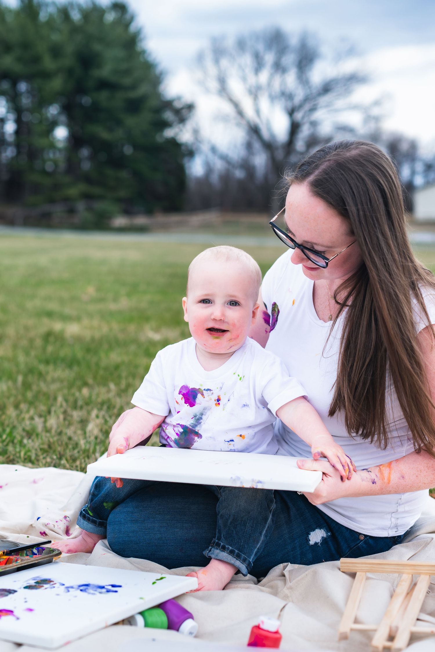 A joyful art session outdoors with paint activities on a blanket in a peaceful park setting.